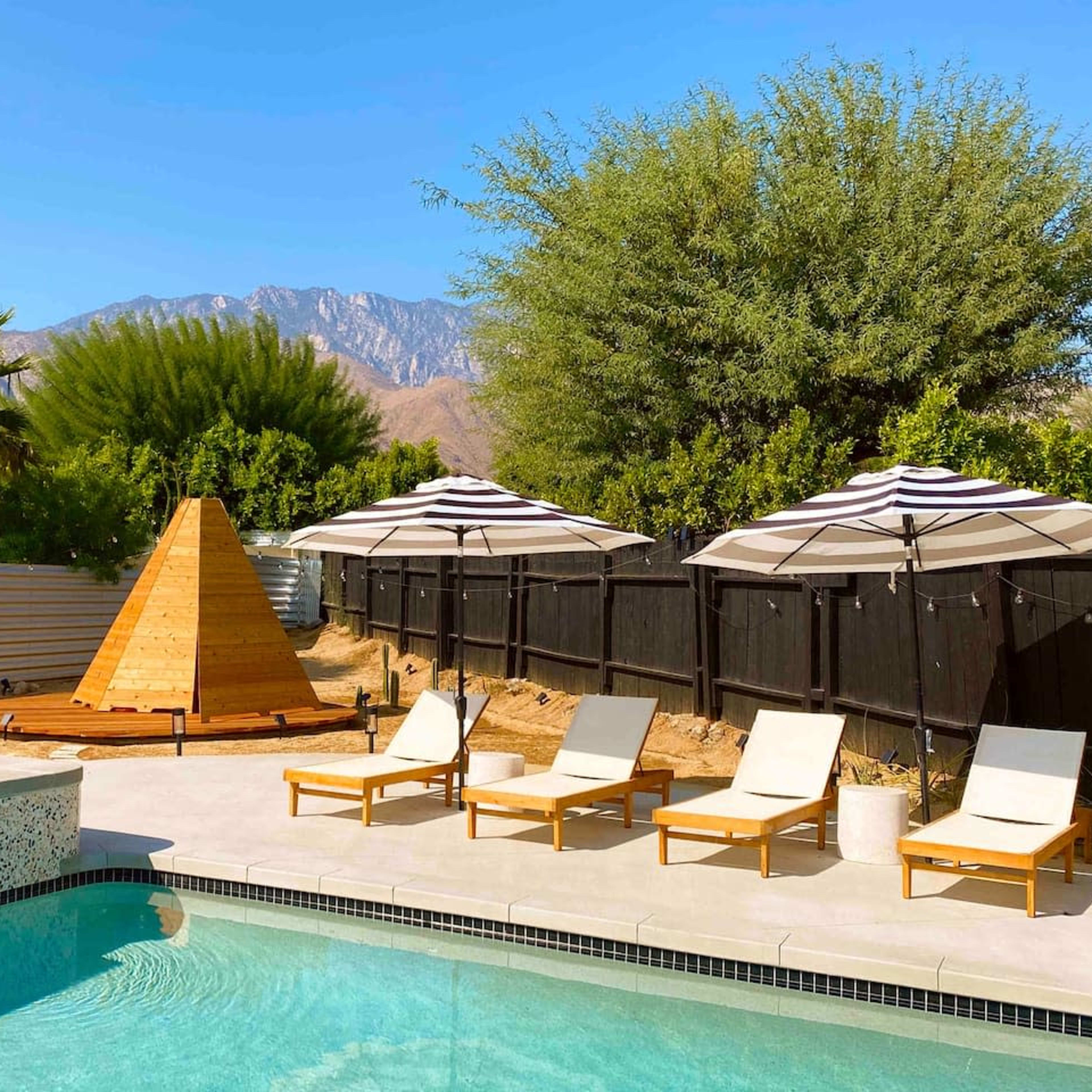 A swimming pool surrounded by lounge chairs and umbrellas, with a wooden pyramid structure and mountains in the background.