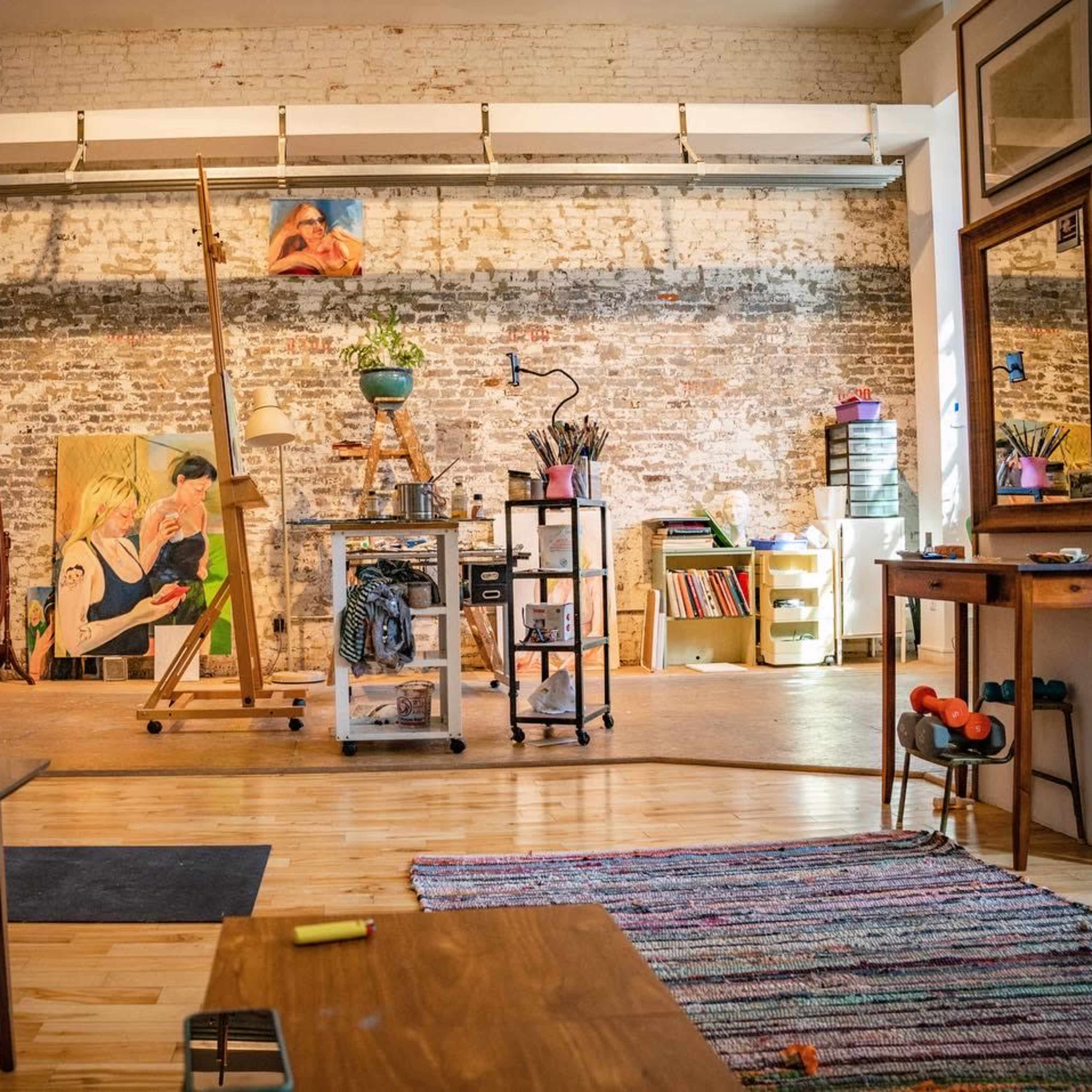 An artist's studio features wooden floors, exposed brick walls, an easel with artwork, various art supplies, and a cozy seating area.