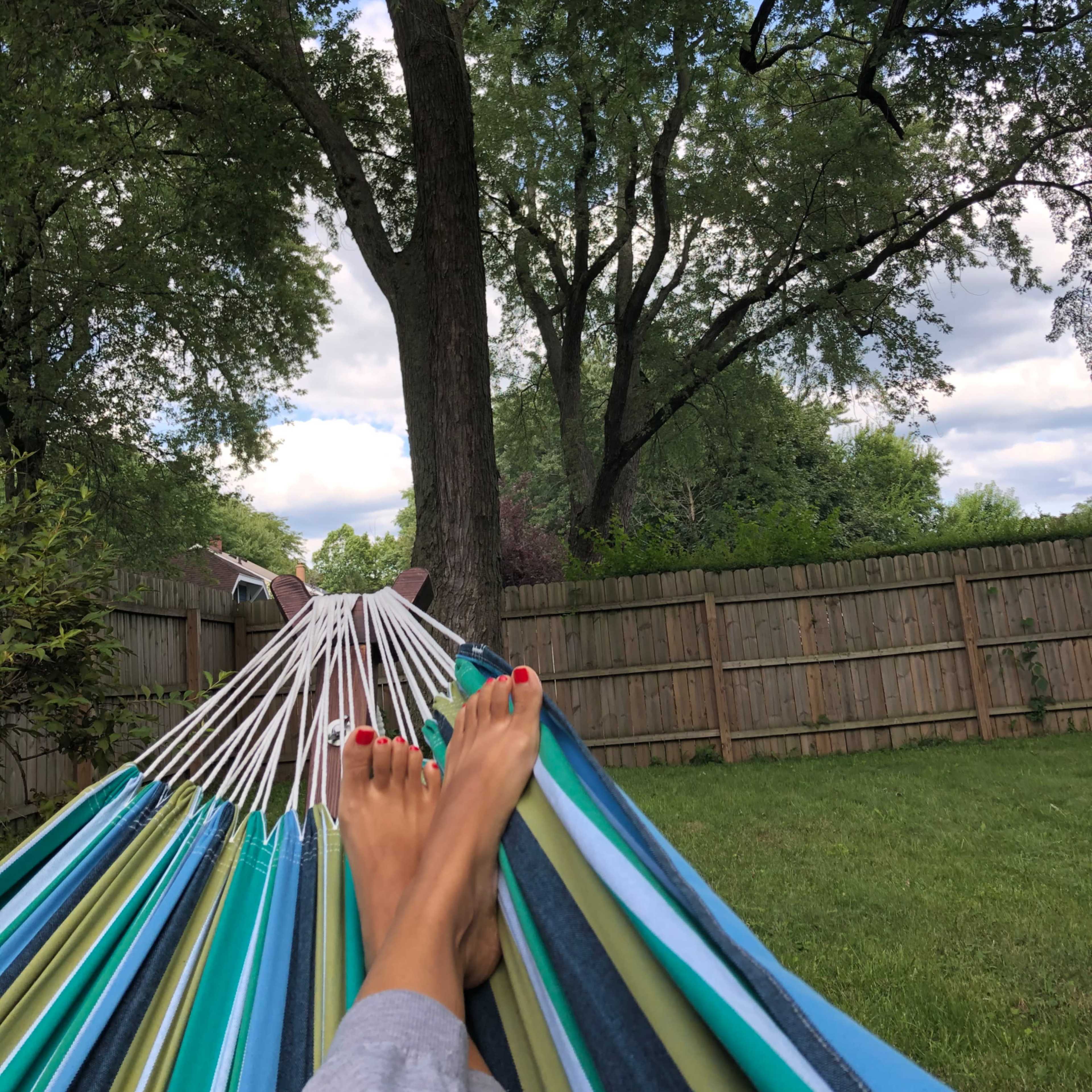 A person relaxes on a colorful hammock in a backyard, with trees and a wooden fence in the background.
