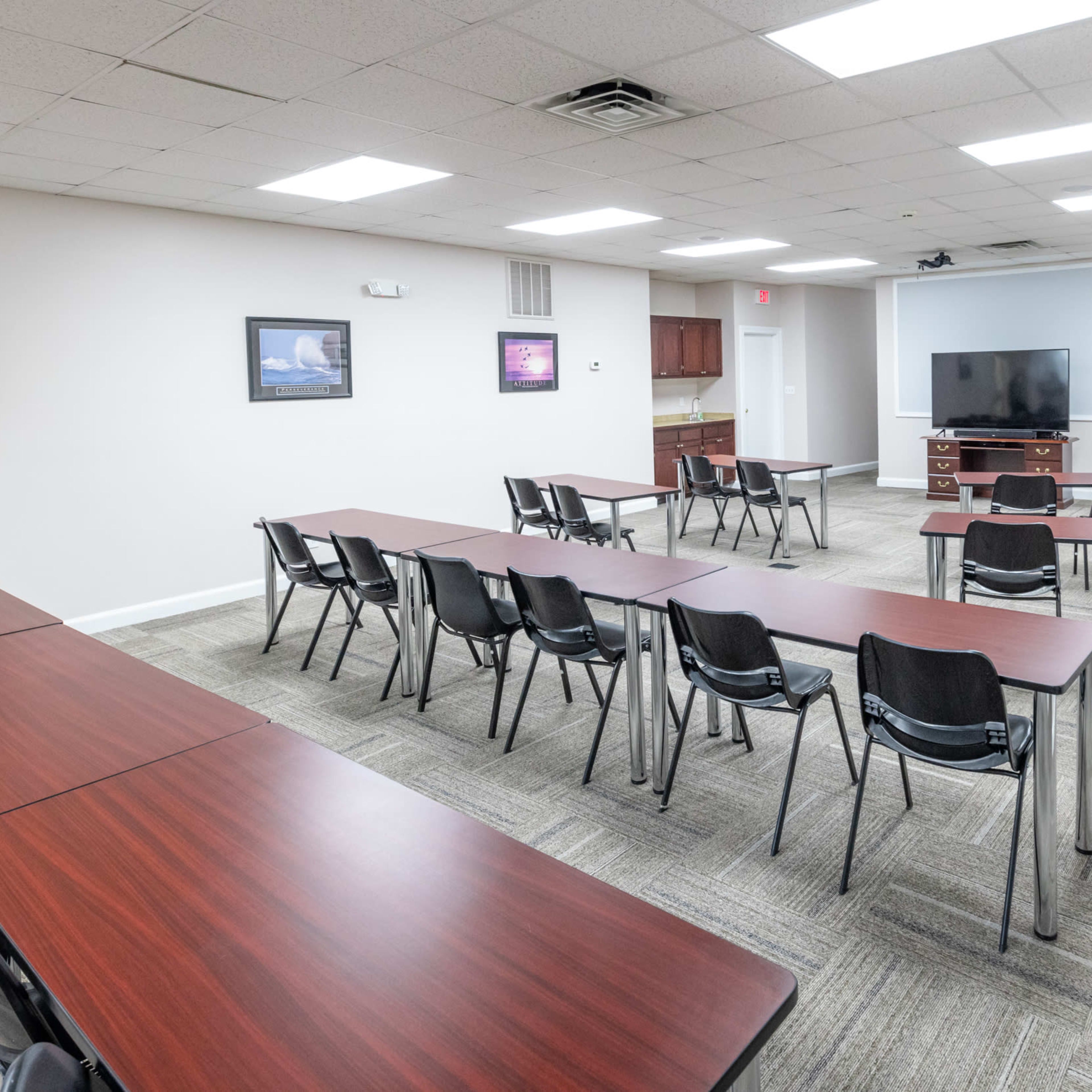 A meeting room with multiple tables arranged in rows and black chairs, along with a television and some framed artwork on the walls.