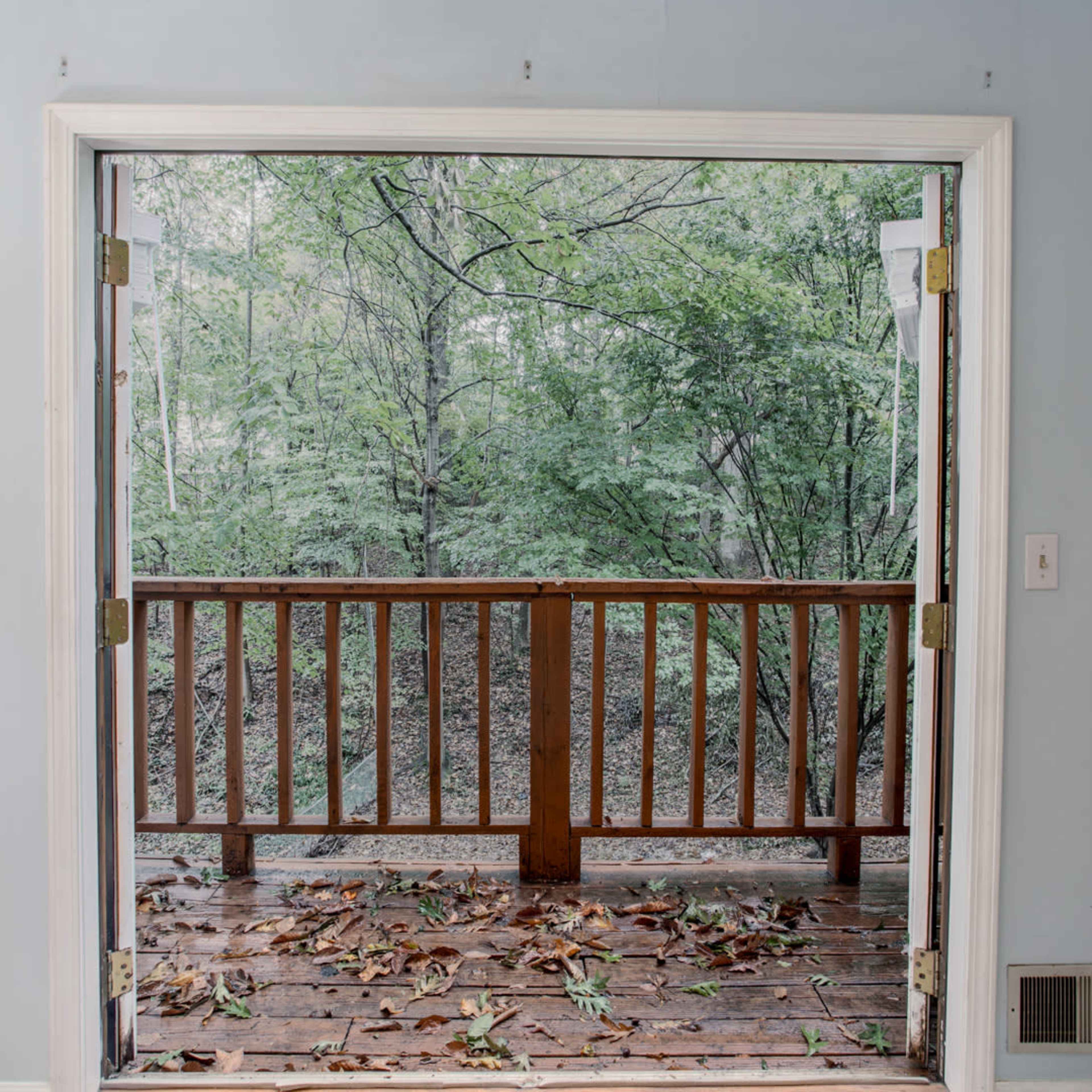 A door opens to a wooden balcony overlooking a tree-filled yard with fallen leaves scattered on the floor.