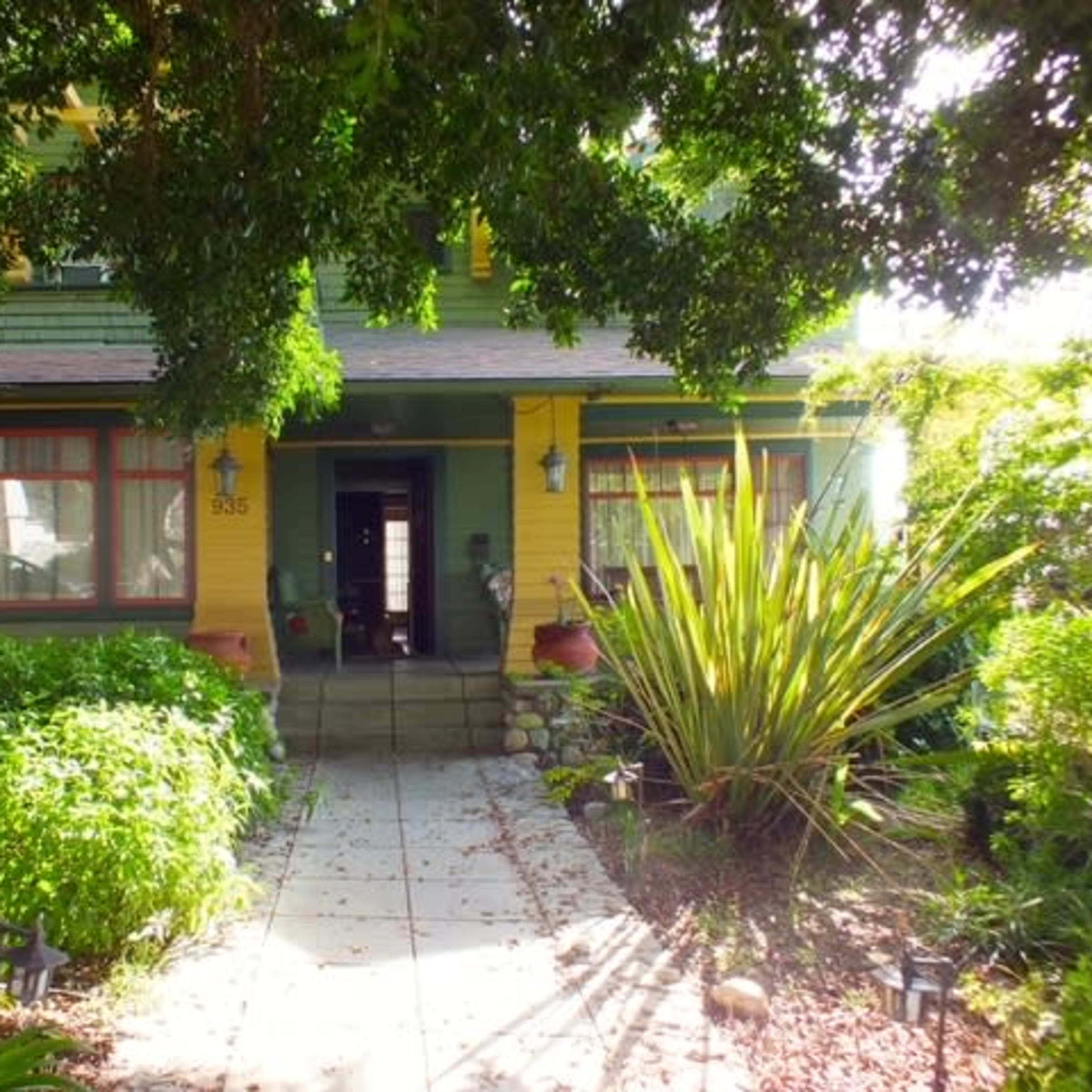 A two-story house with yellow and green exterior paint, surrounded by lush greenery and a stone pathway leading to the front door.