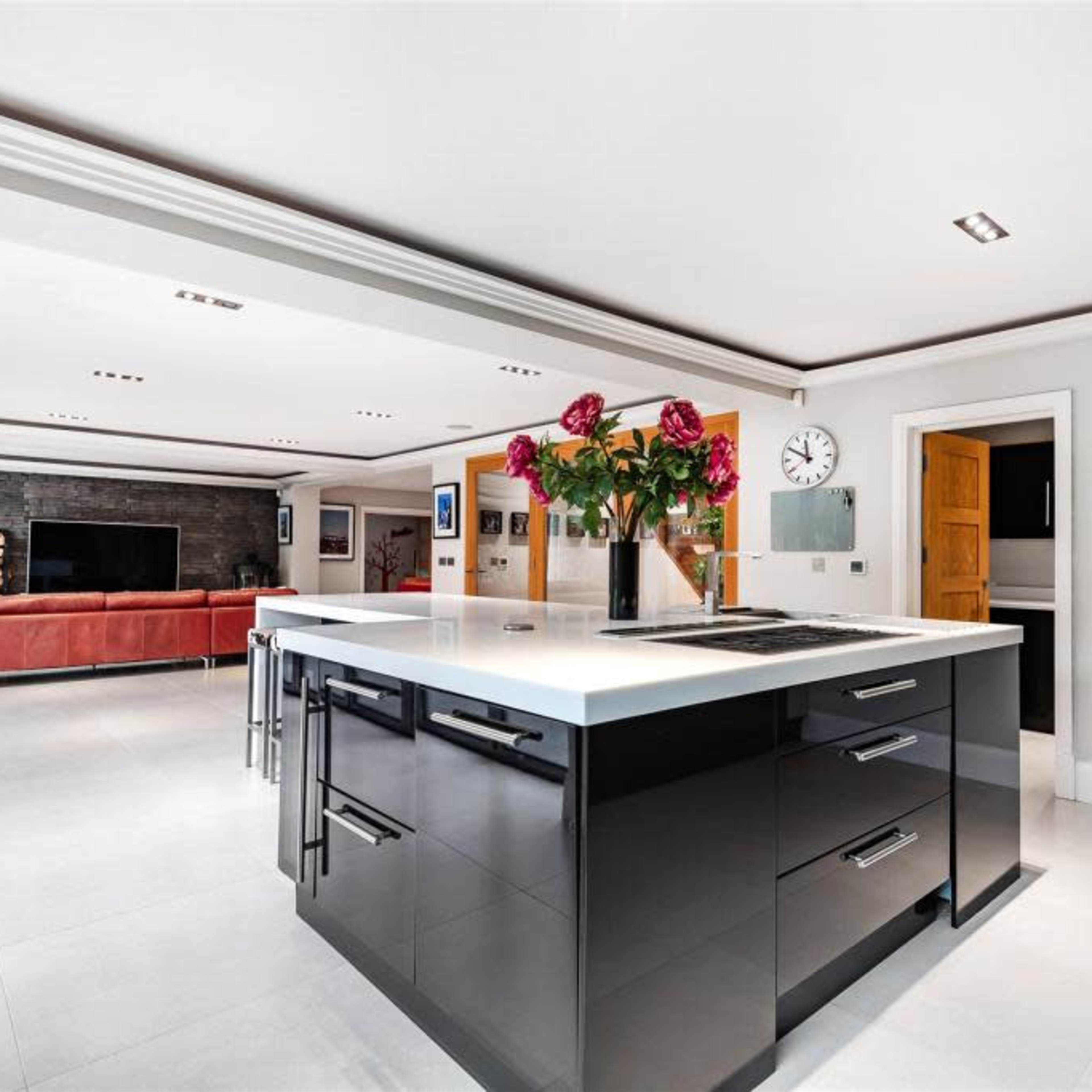 A modern kitchen features a central island with a cooktop and flowers, adjacent to a living area with a large sofa and a television.