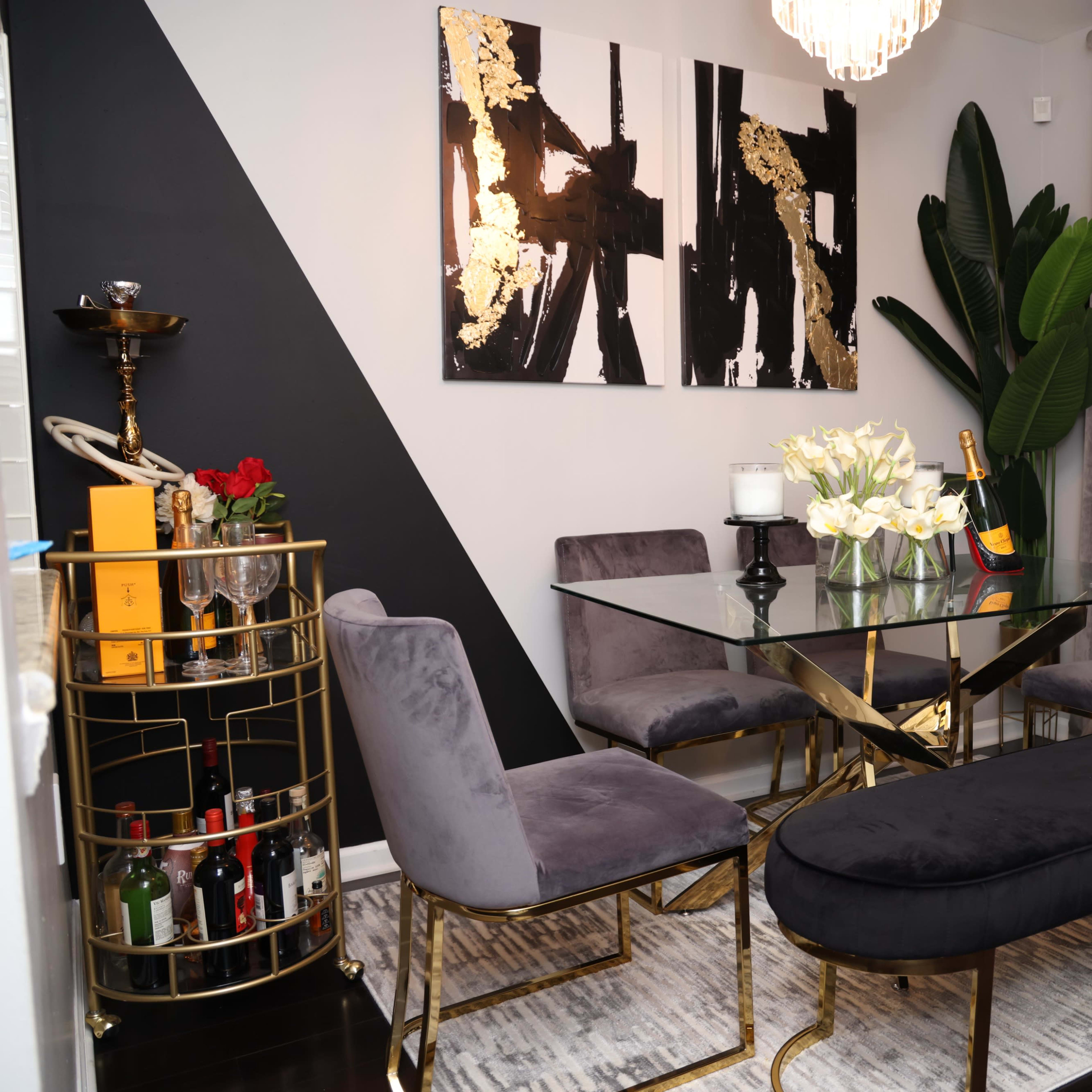 A modern dining area features a glass table surrounded by four gray chairs, with a gold-accented bar cart and two abstract paintings on the wall.