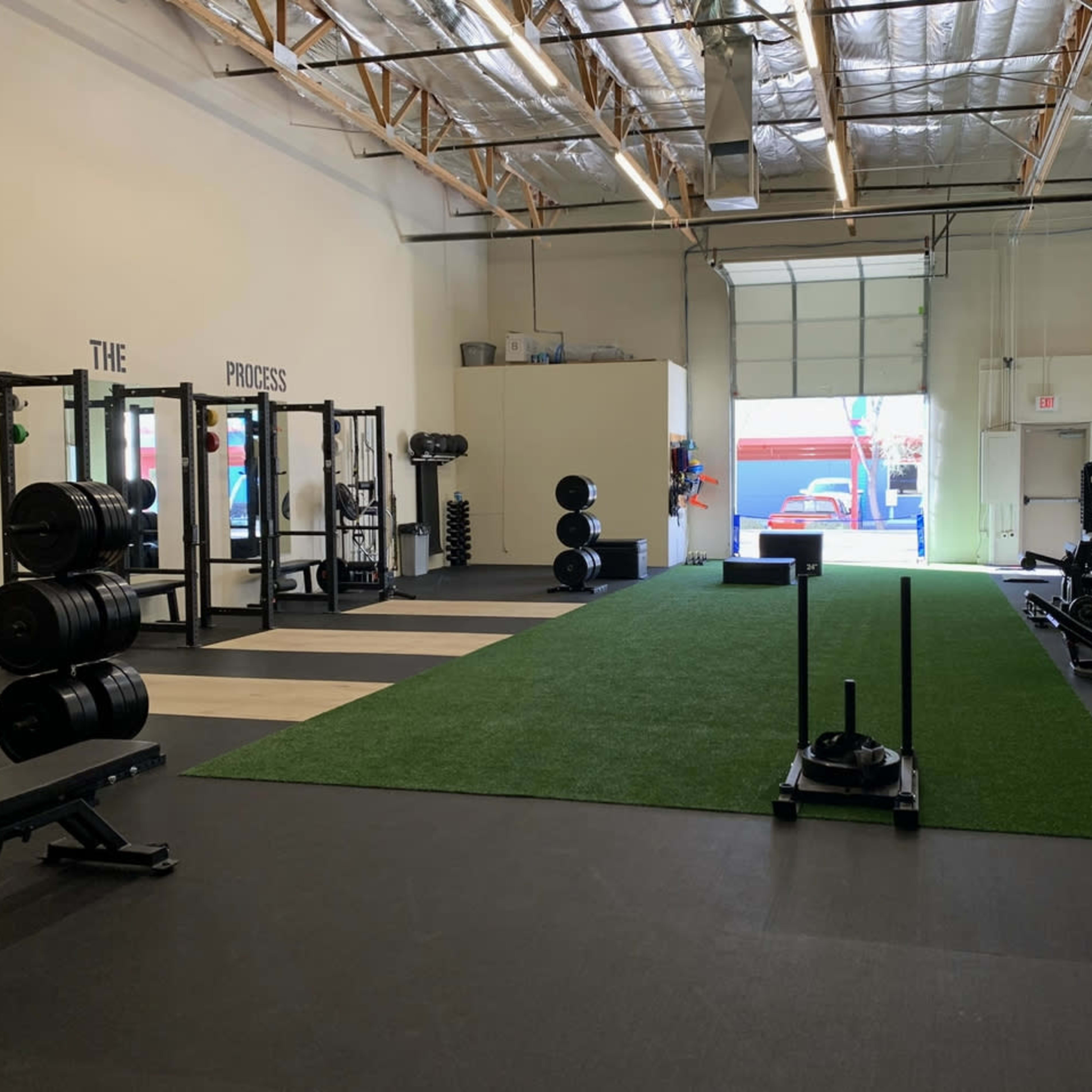 A fitness facility with a turf area, various weightlifting equipment, and an open garage door leading outside.