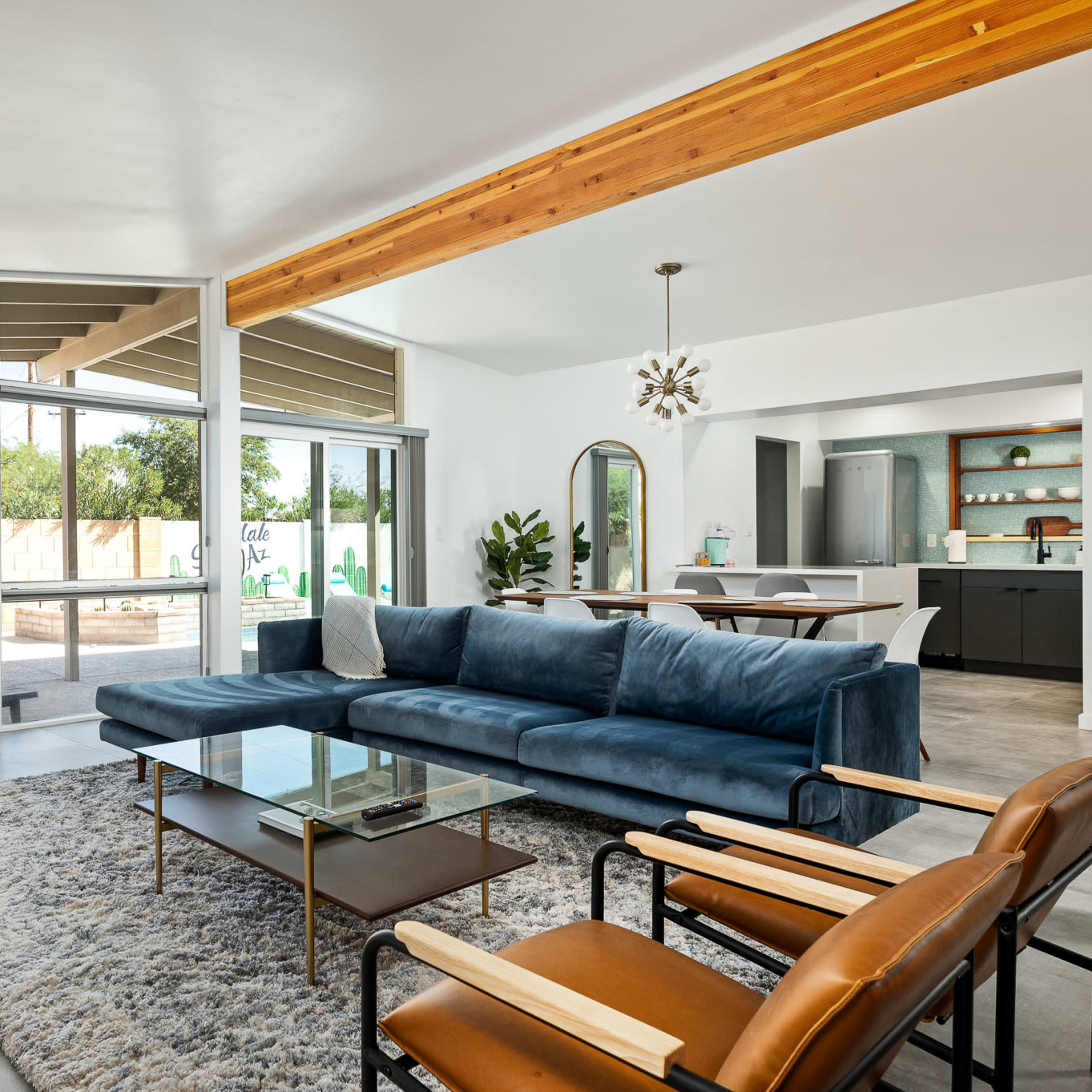 A modern living room featuring a blue sectional sofa, a glass coffee table, and a unique fireplace, with large windows overlooking a backyard.