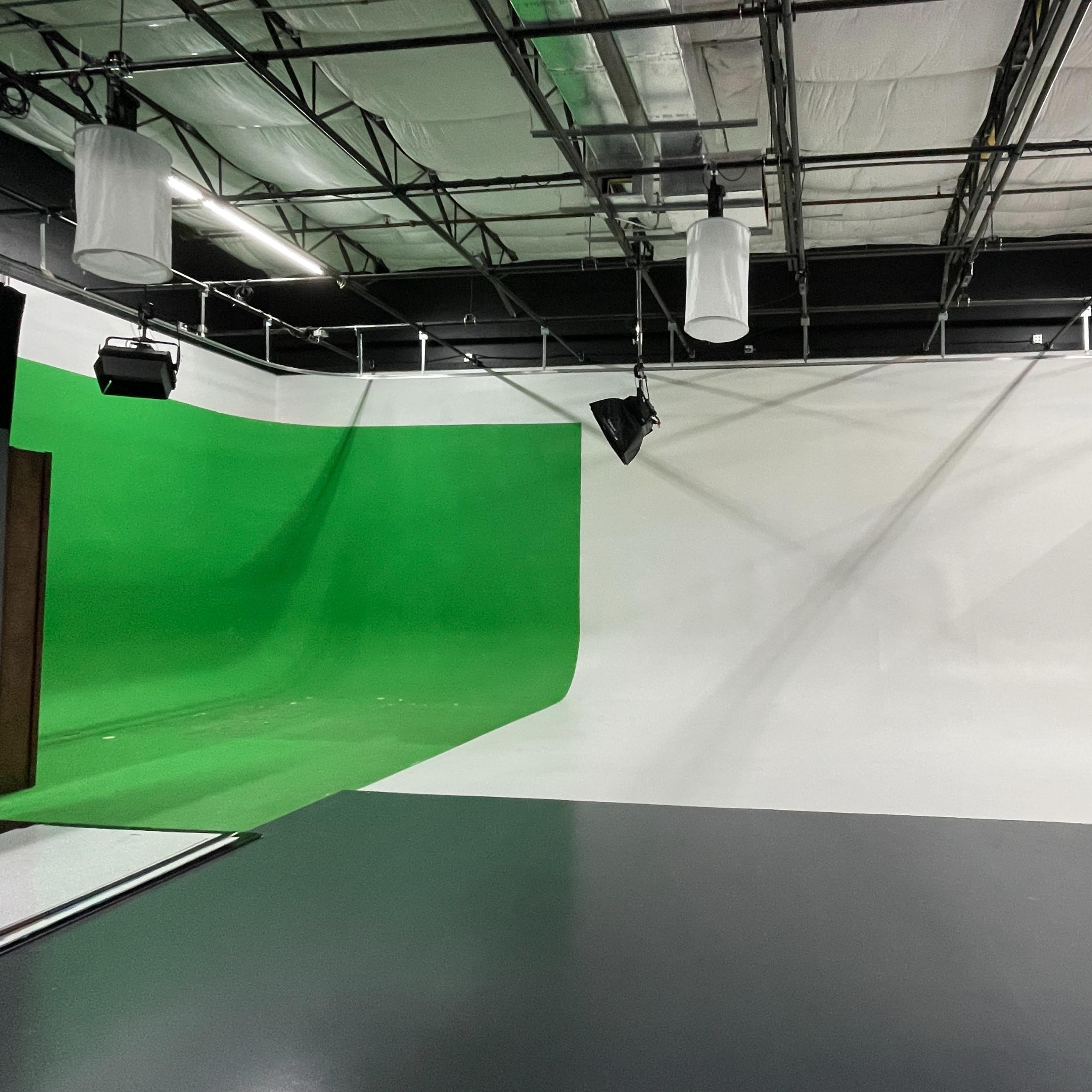 A partially empty studio space with a green screen on one side and a white background on the other, under a ceiling with hanging lights.