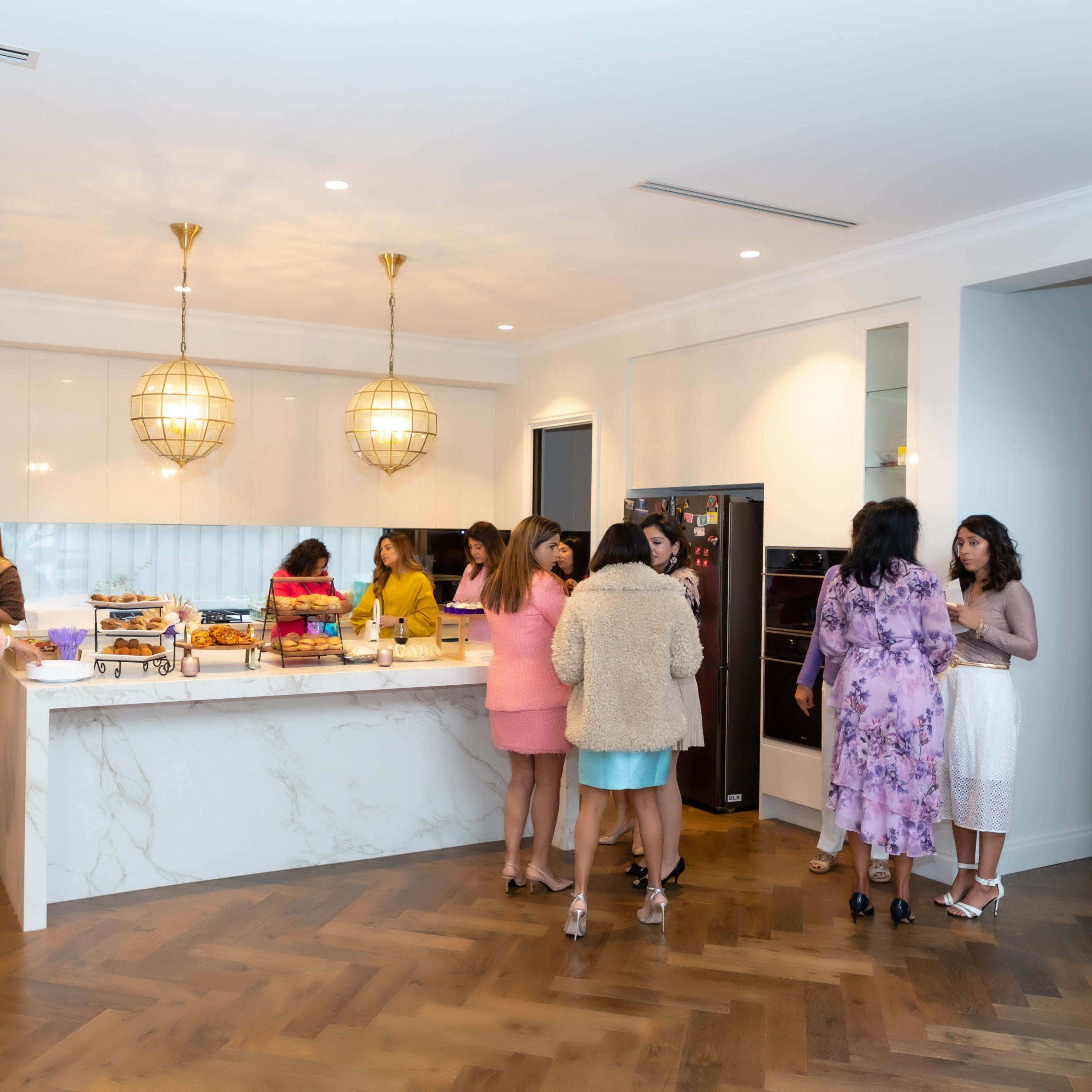 A group of women gathers in a modern kitchen with a large marble island, preparing food and drinks.