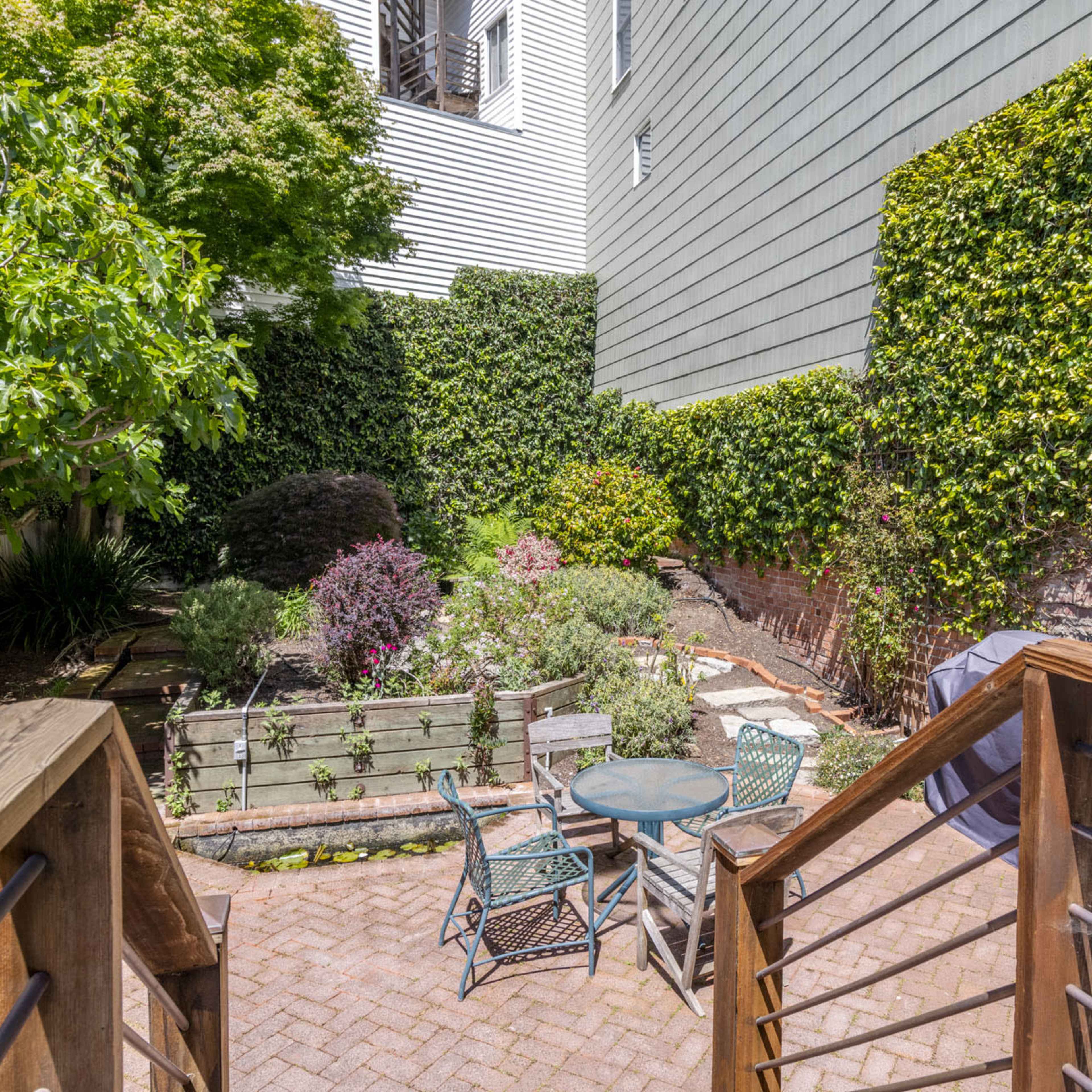 A garden area with a stone patio, wooden steps leading down, a circular table with chairs, and lush greenery along the walls.