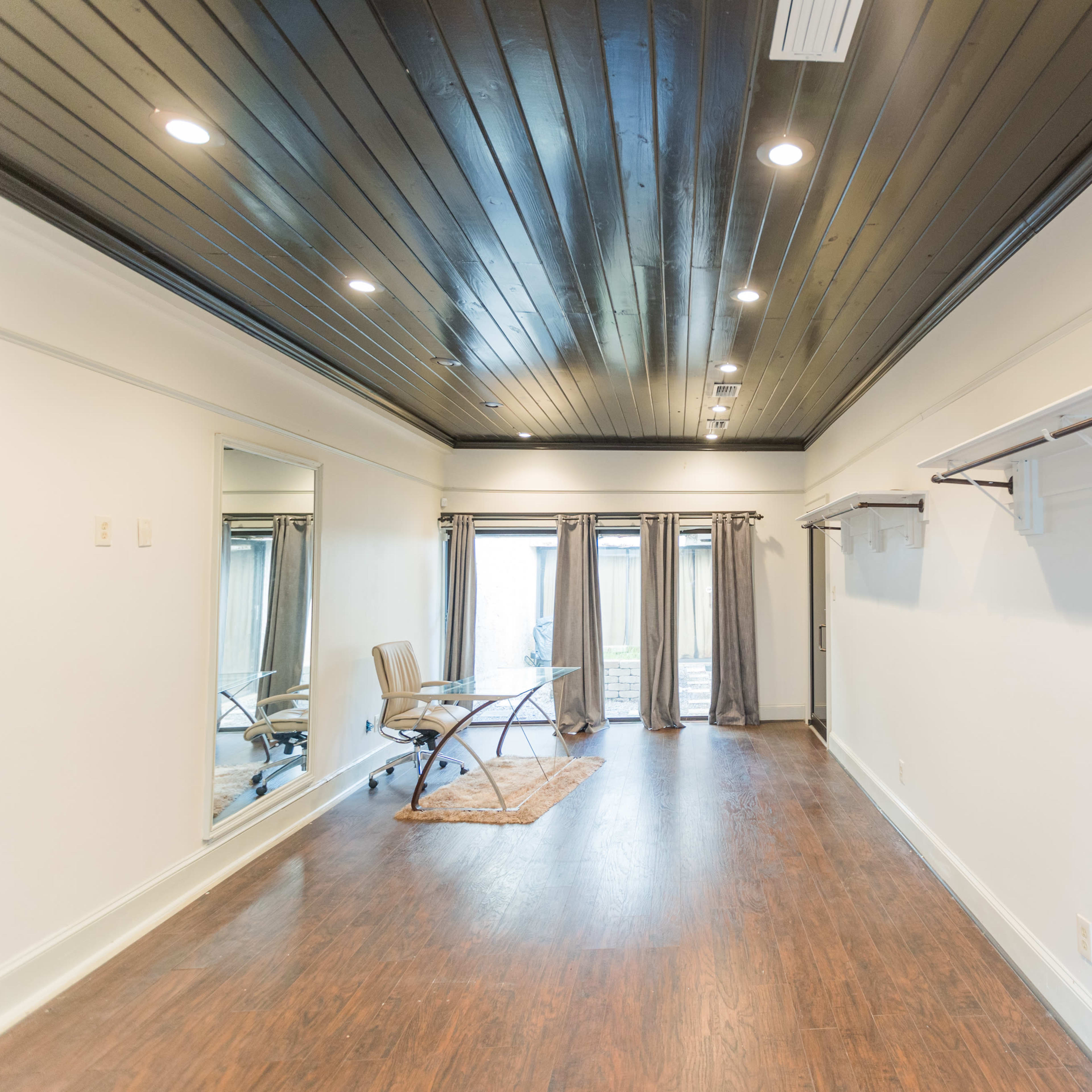 A spacious room with a dark wooden ceiling, a large mirror on one wall, and a chair positioned near a window with sheer curtains.