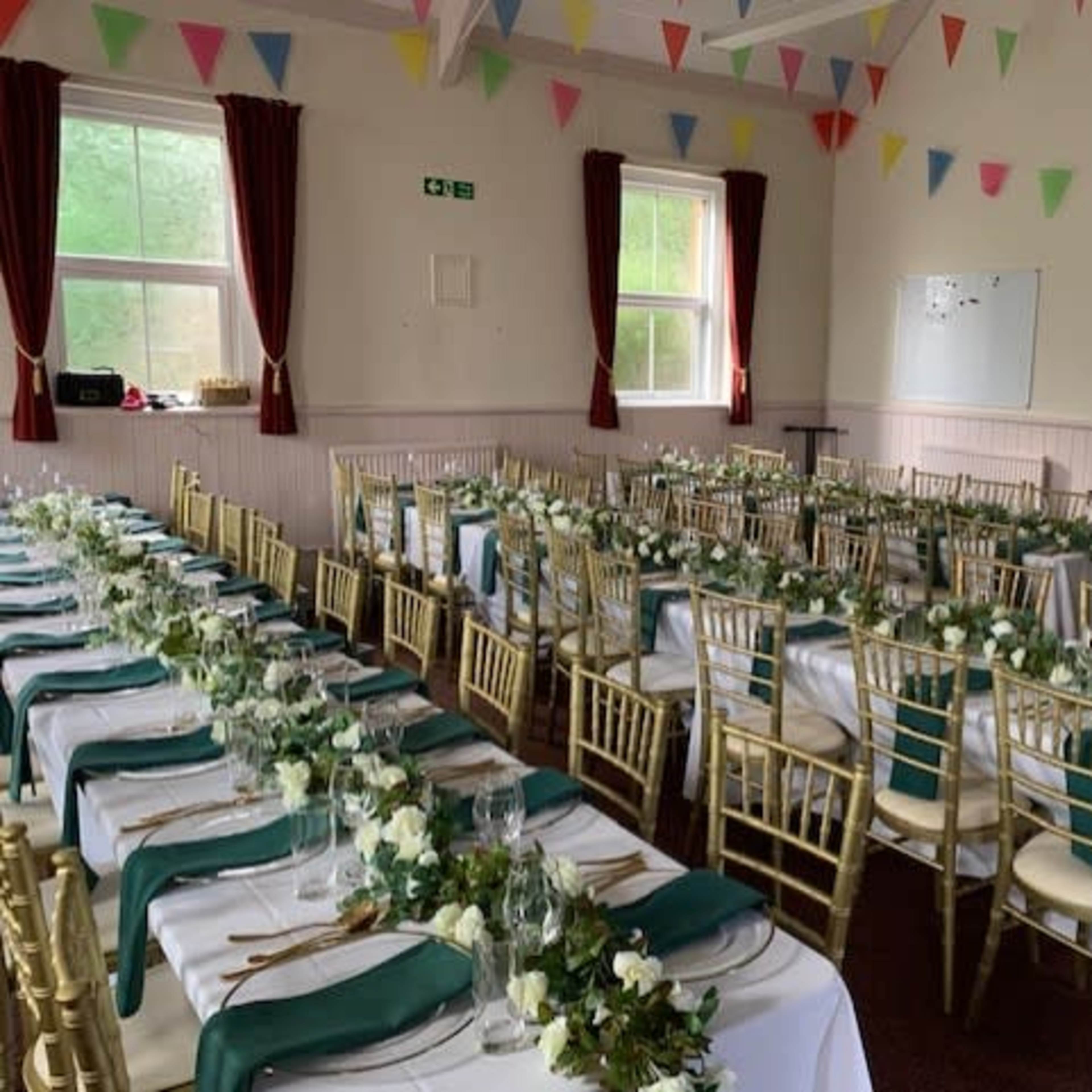 A banquet hall arranged with multiple long tables covered in white cloths, adorned with greenery and floral centerpieces, and decorated with colorful bunting overhead.