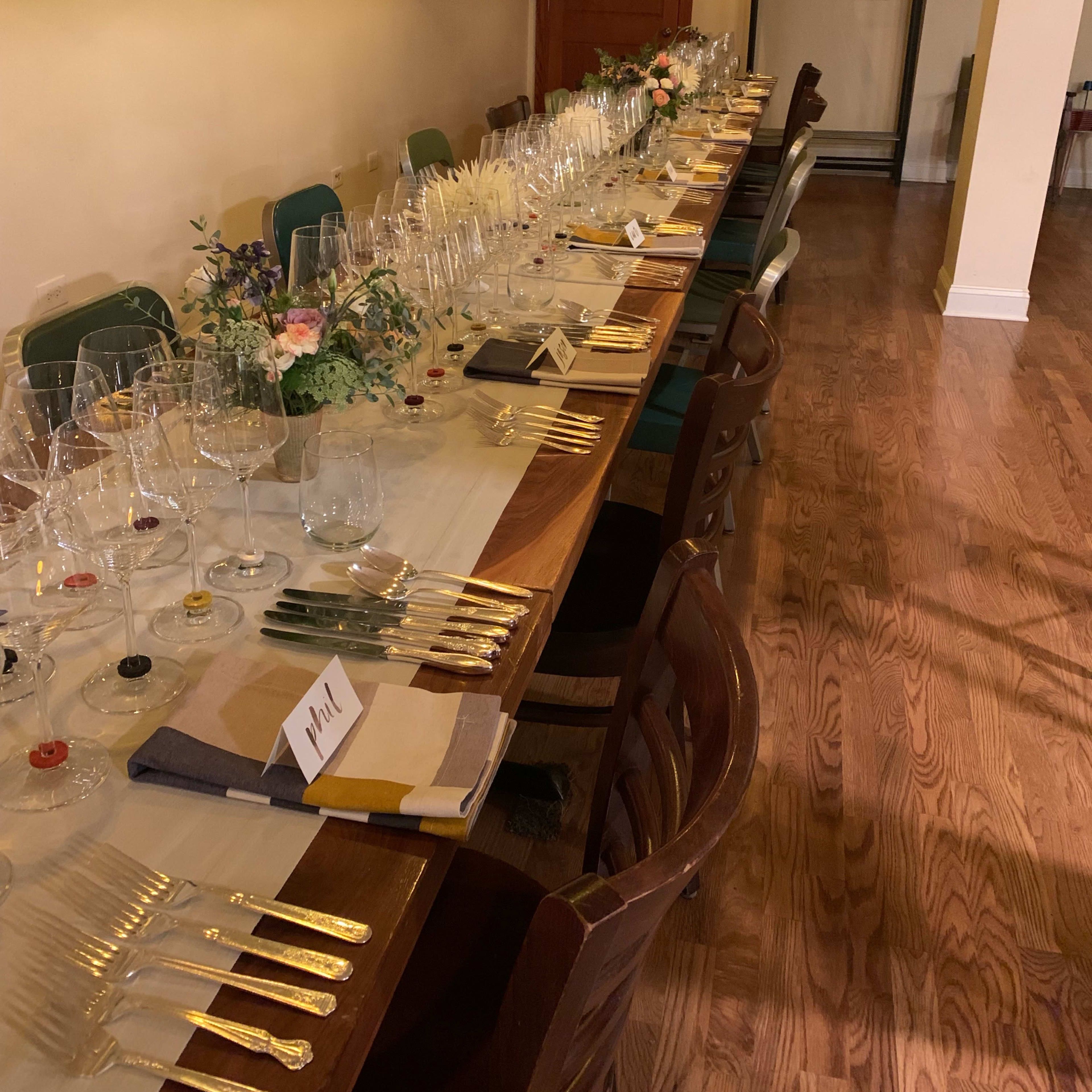 A long wooden dining table is set with glassware and silverware in a well-lit room, with framed mirrors on the wall and floral arrangements as centerpieces.