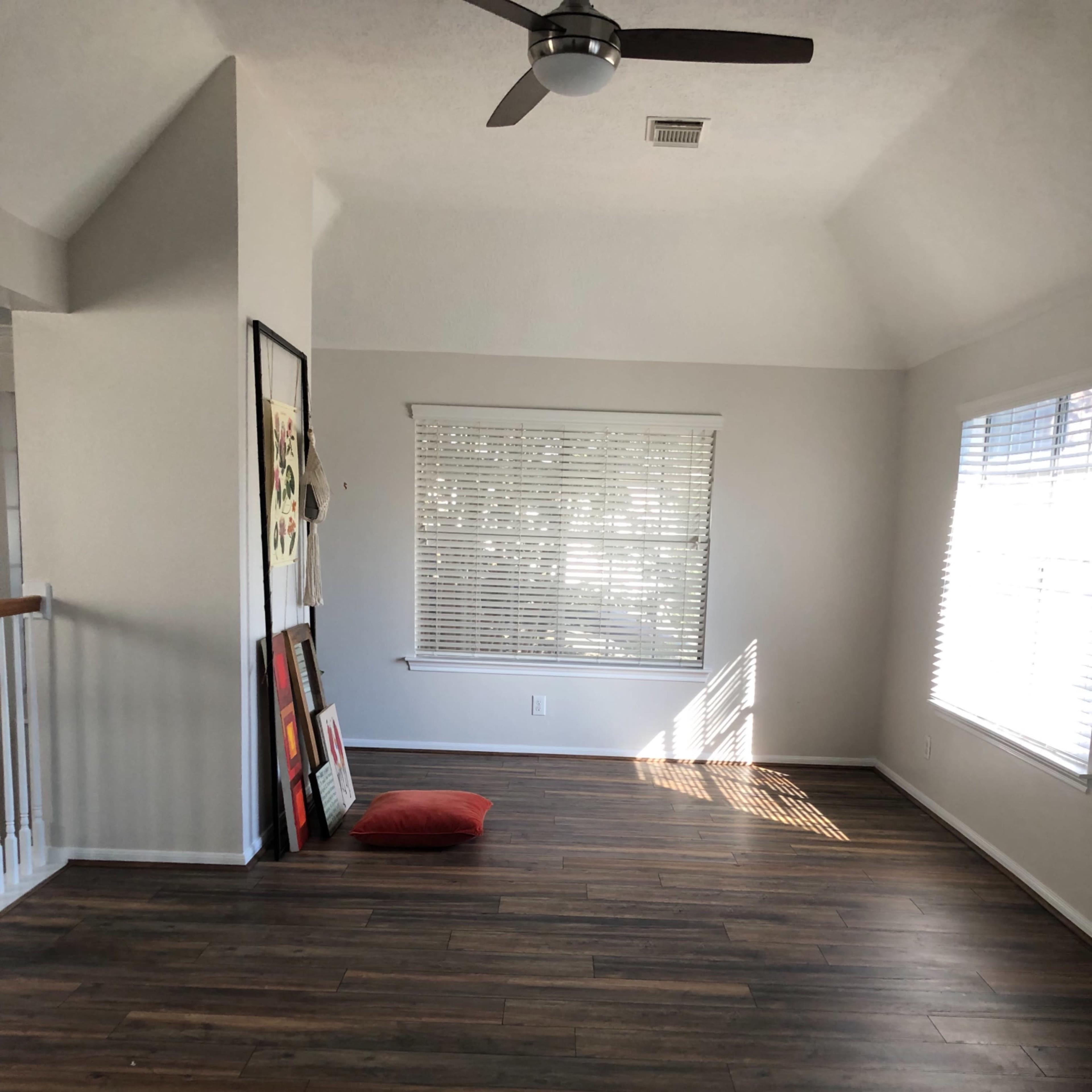 An empty room with wood-like floors, a ceiling fan, large windows with blinds, and a red cushion on the floor near a wall with framed artwork.