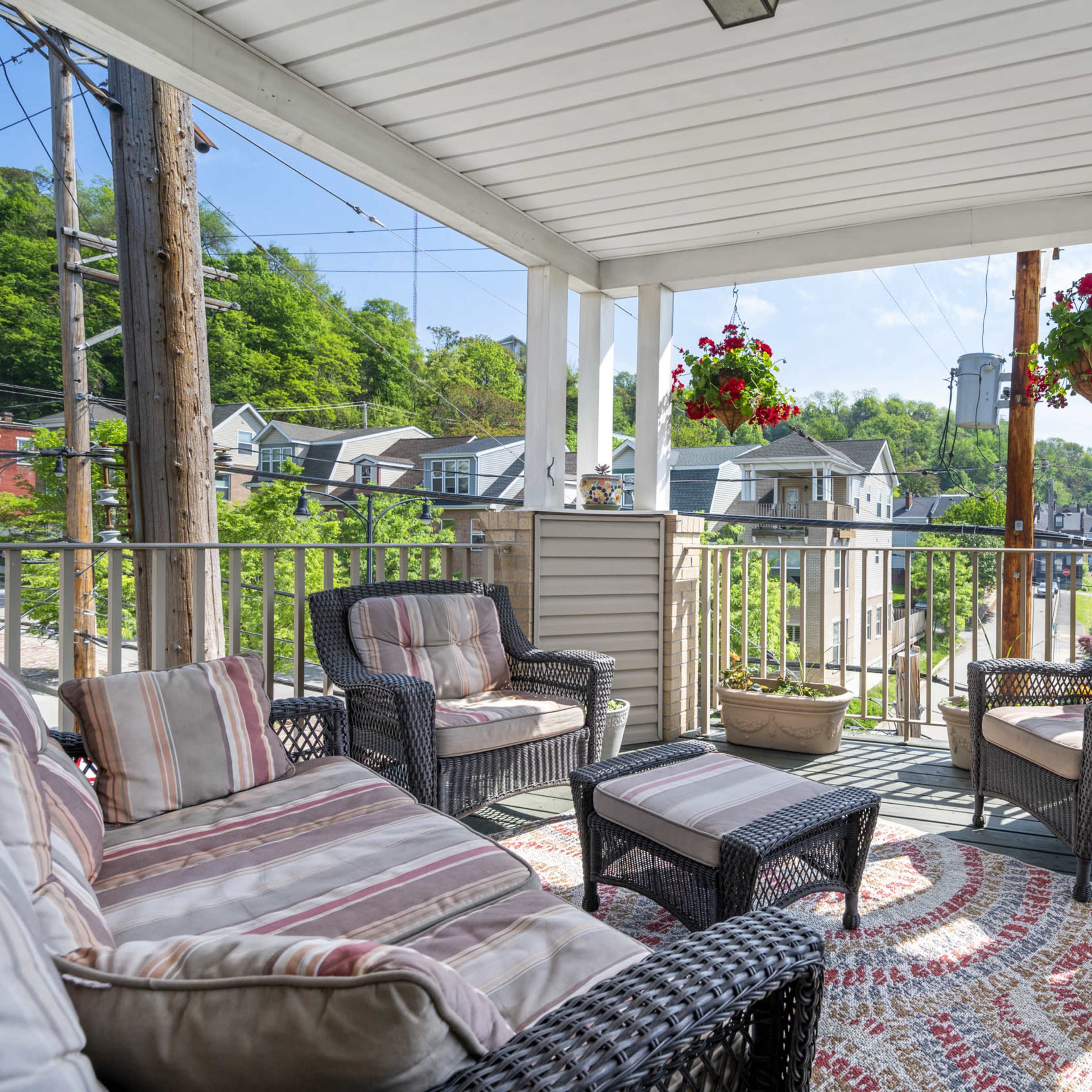 A covered porch features wicker seating, a decorative rug, and hanging flower baskets, overlooking a neighborhood with trees and houses in the background.