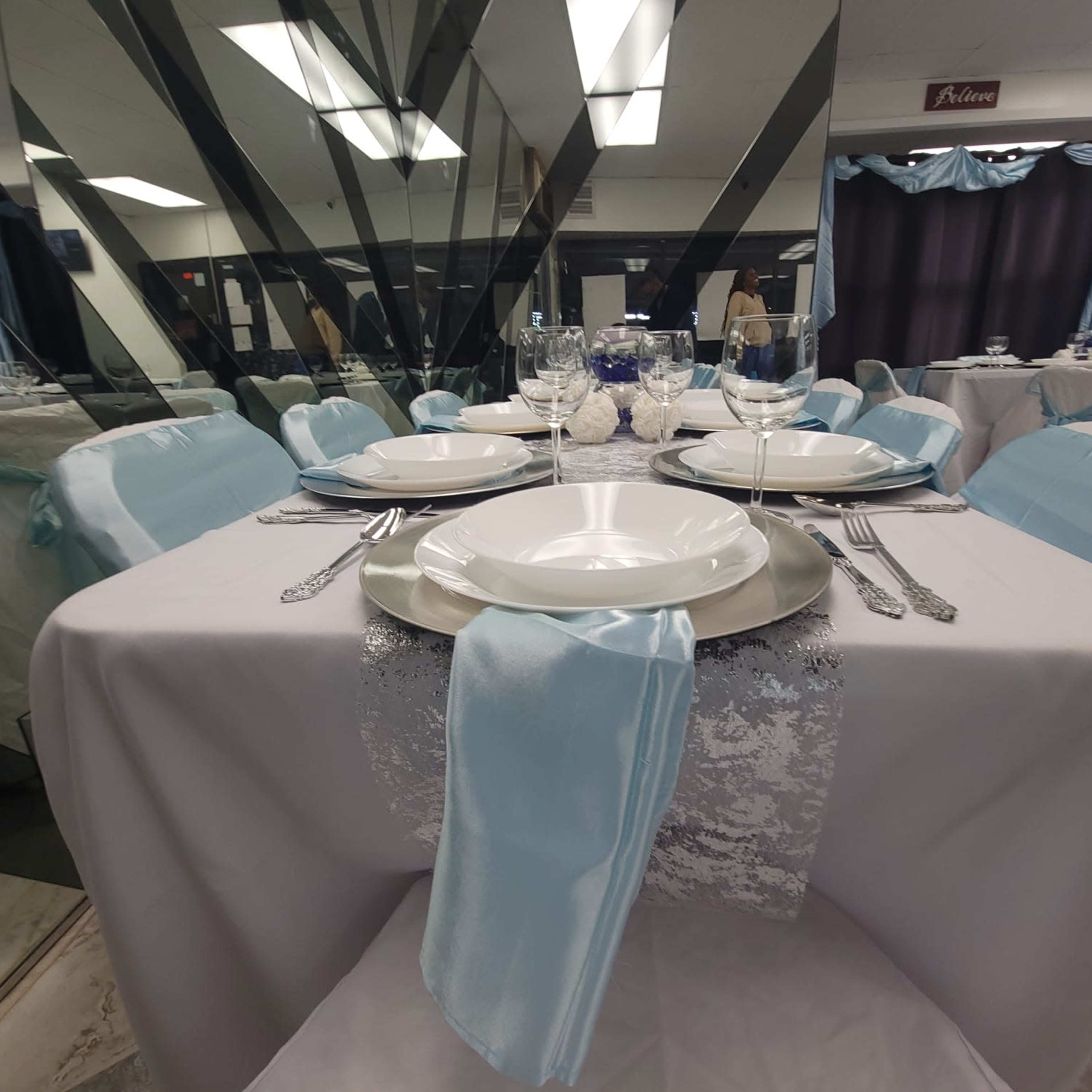 A banquet table is elegantly set with plates, glassware, and a light blue table runner, surrounded by decorative mirrors.