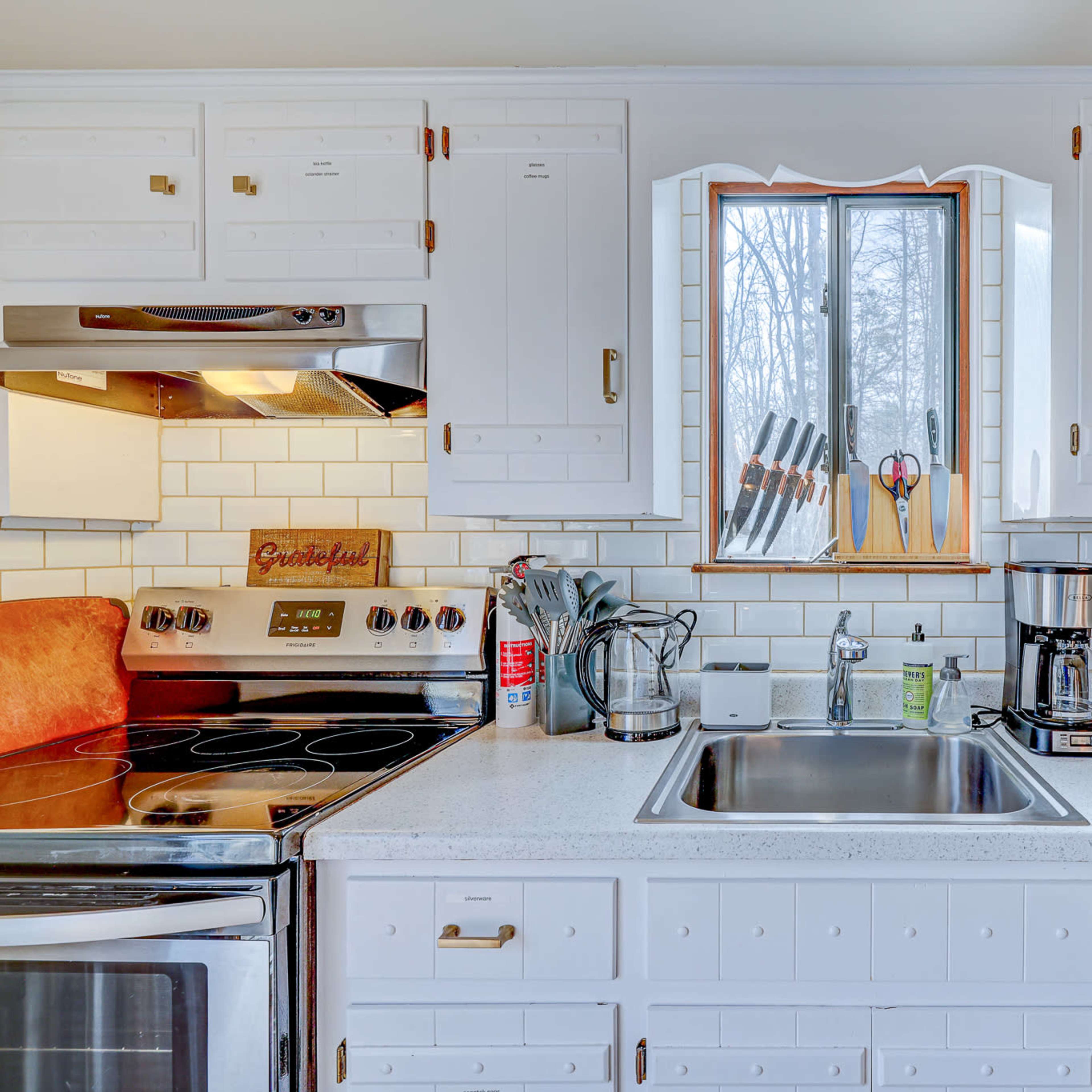 A kitchen features white cabinets, a sink beneath a window, an electric stove, a coffee maker, and a countertop toaster oven.