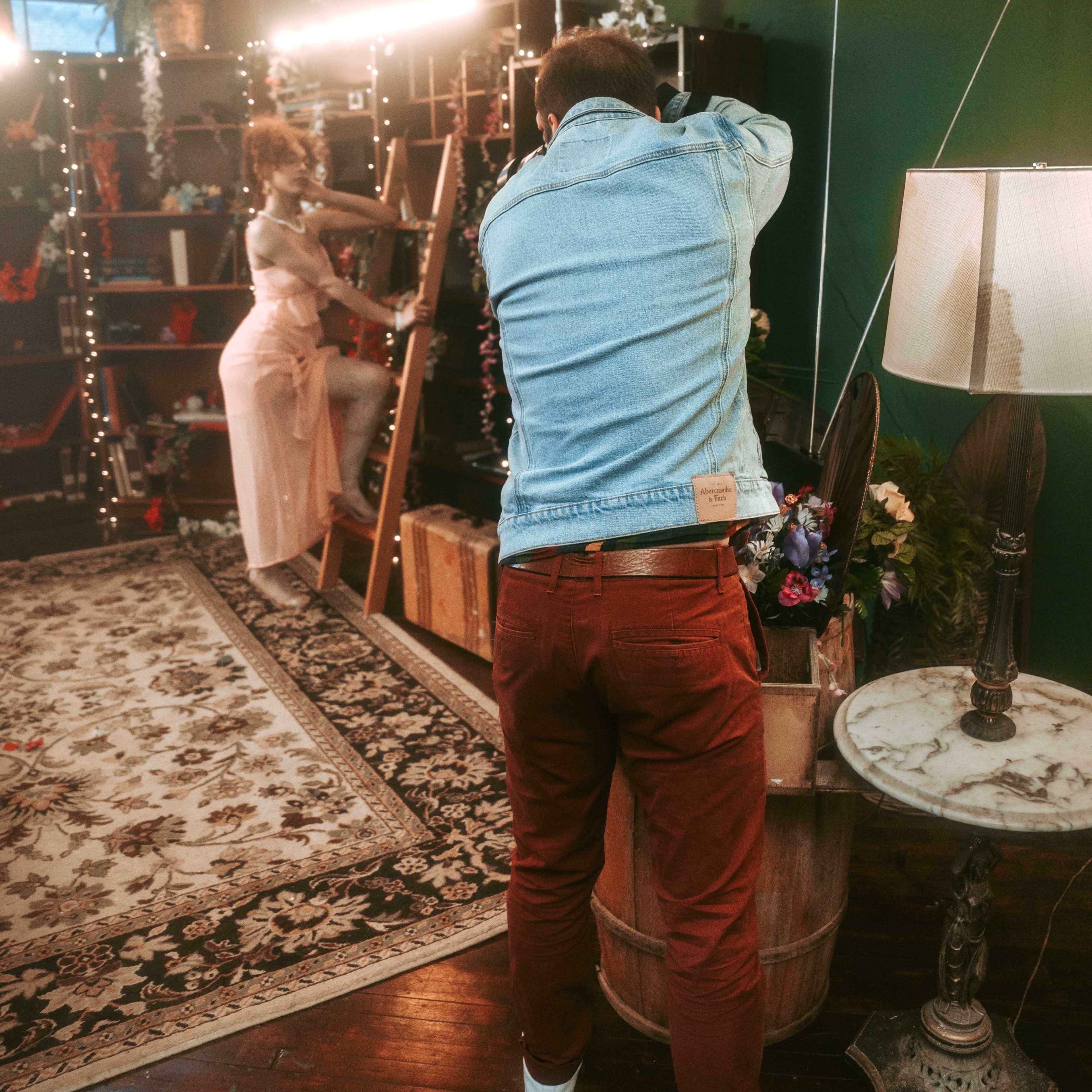 A photographer captures a model posing on a ladder in a decorated studio with greenery and props.
