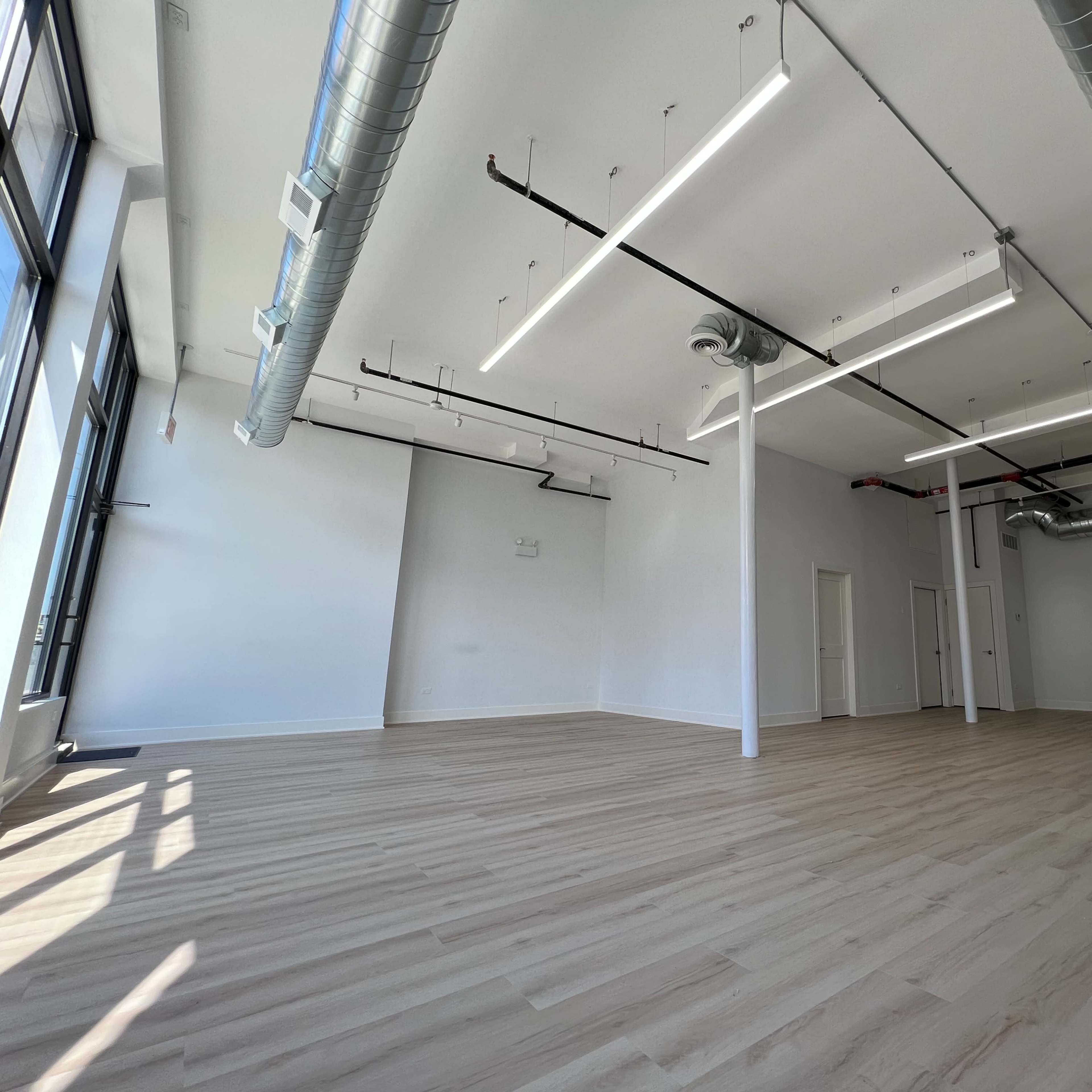 A spacious, empty room with large windows, polished wooden floors, and exposed ductwork on the ceiling.