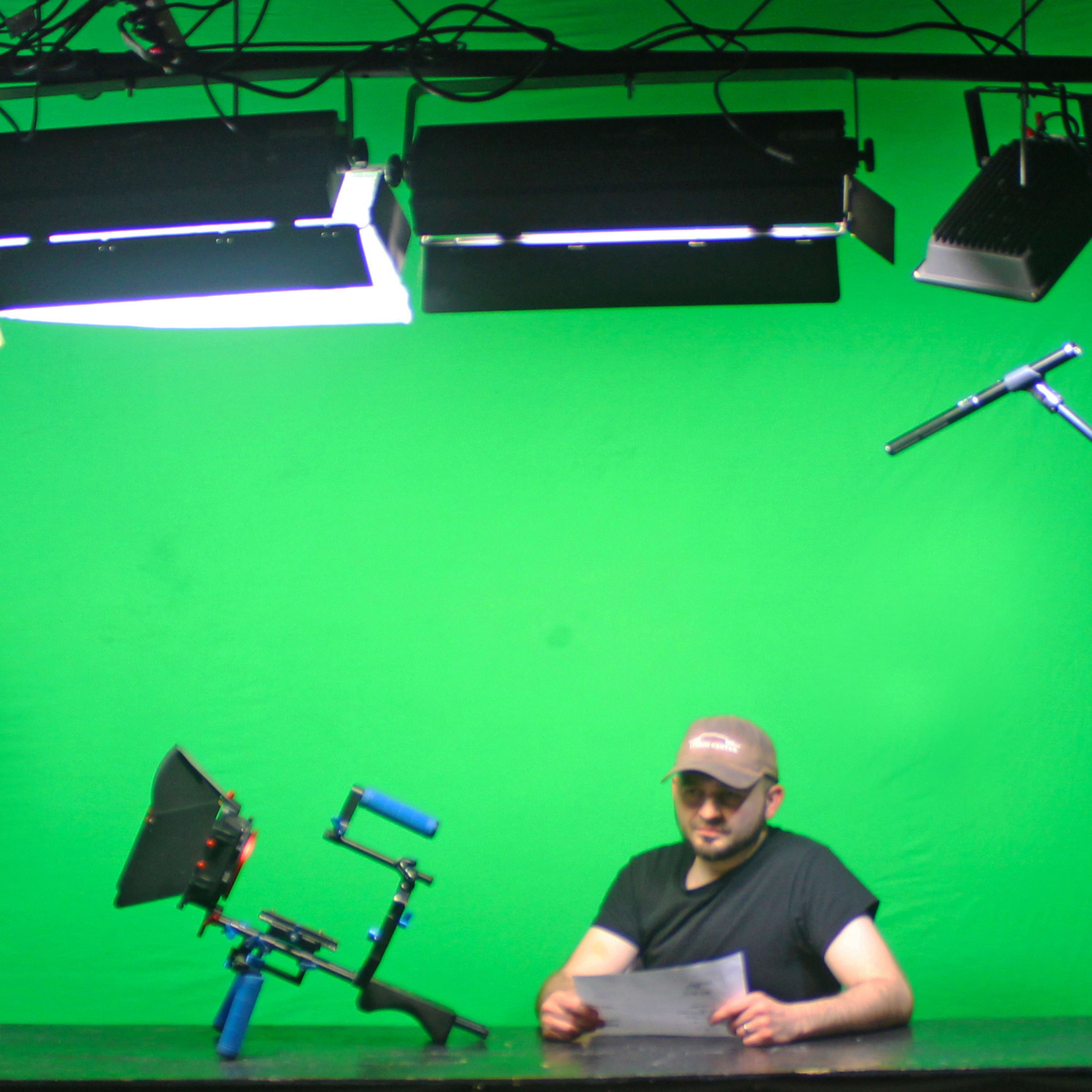 A person sits at a table in front of a green screen, holding a paper, with studio lights and a microphone positioned above them.