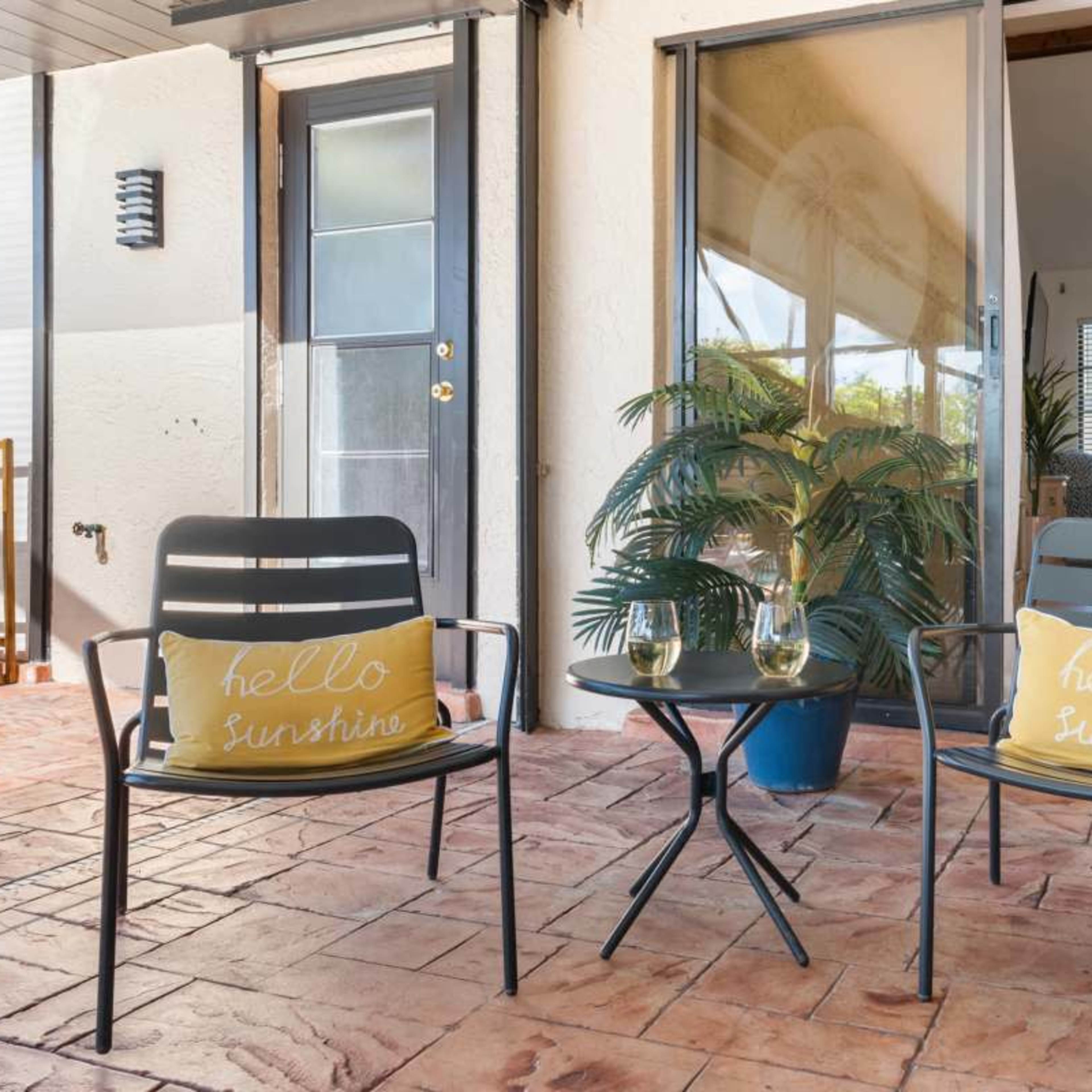 Two chairs with yellow pillows are placed beside a small table on a patio with a potted plant in the background.