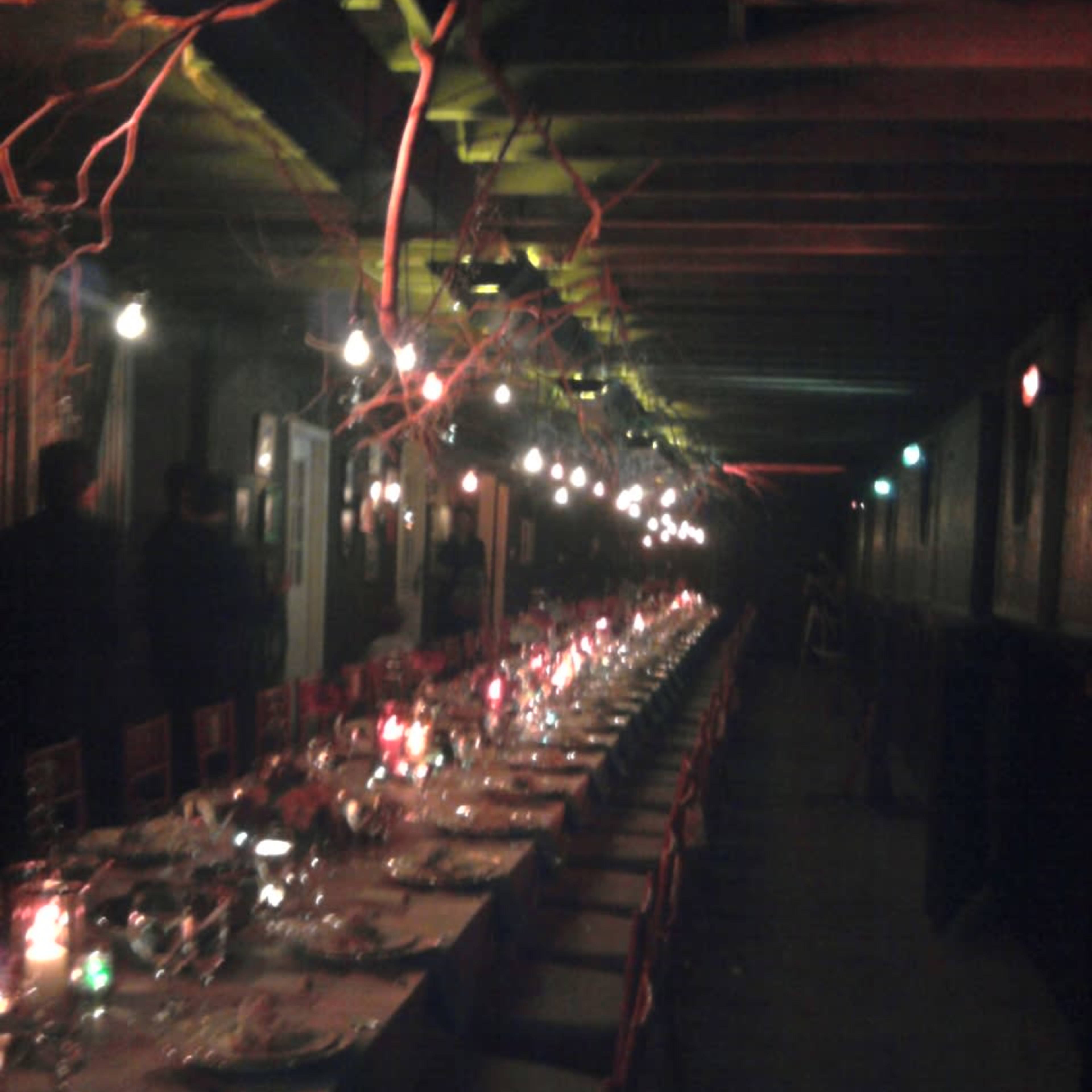 A long dining table is set with multiple place settings and illuminated by hanging lights in a dimly lit room with wooden beams.