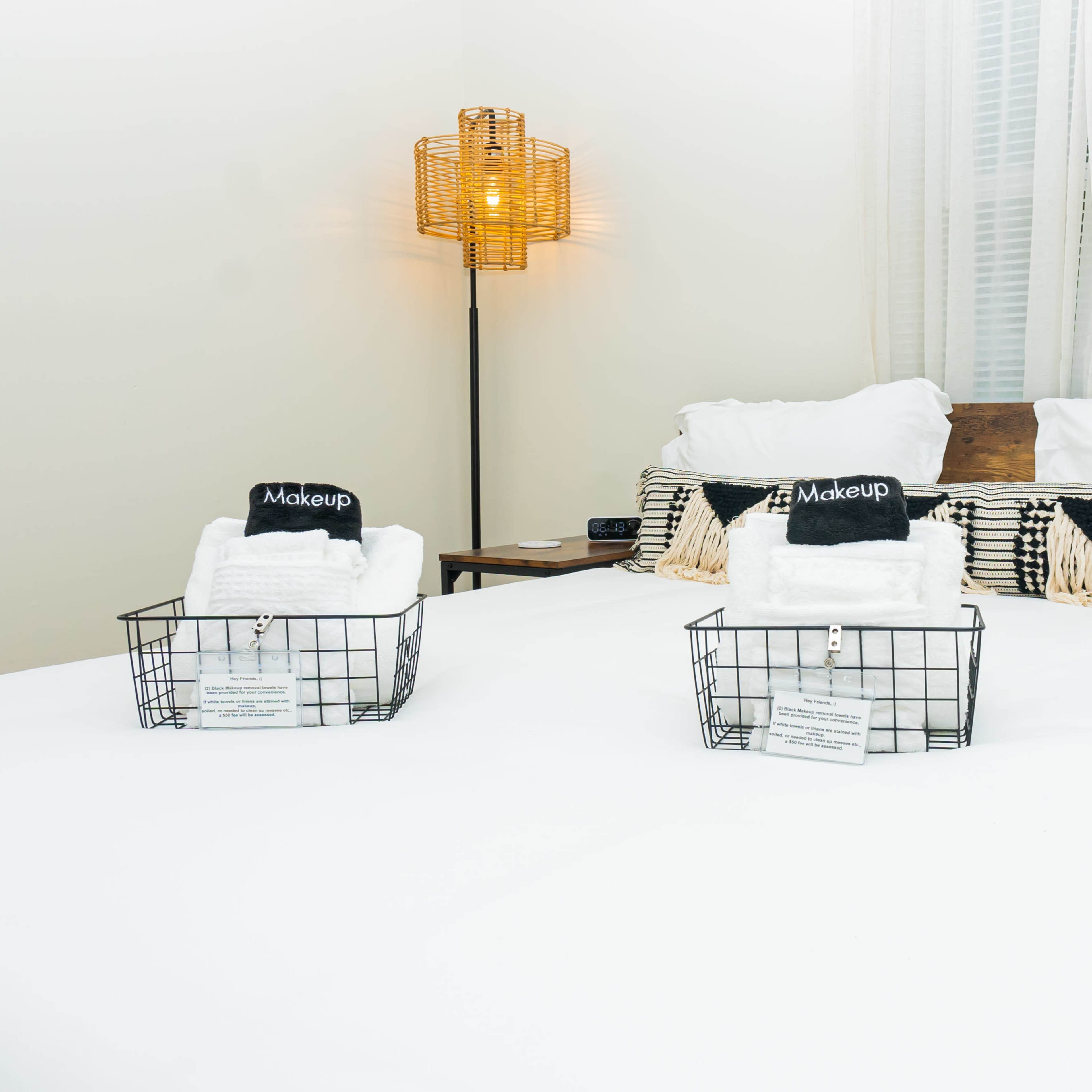 A neatly made bed with two wire baskets of rolled towels sits in a well-lit bedroom.