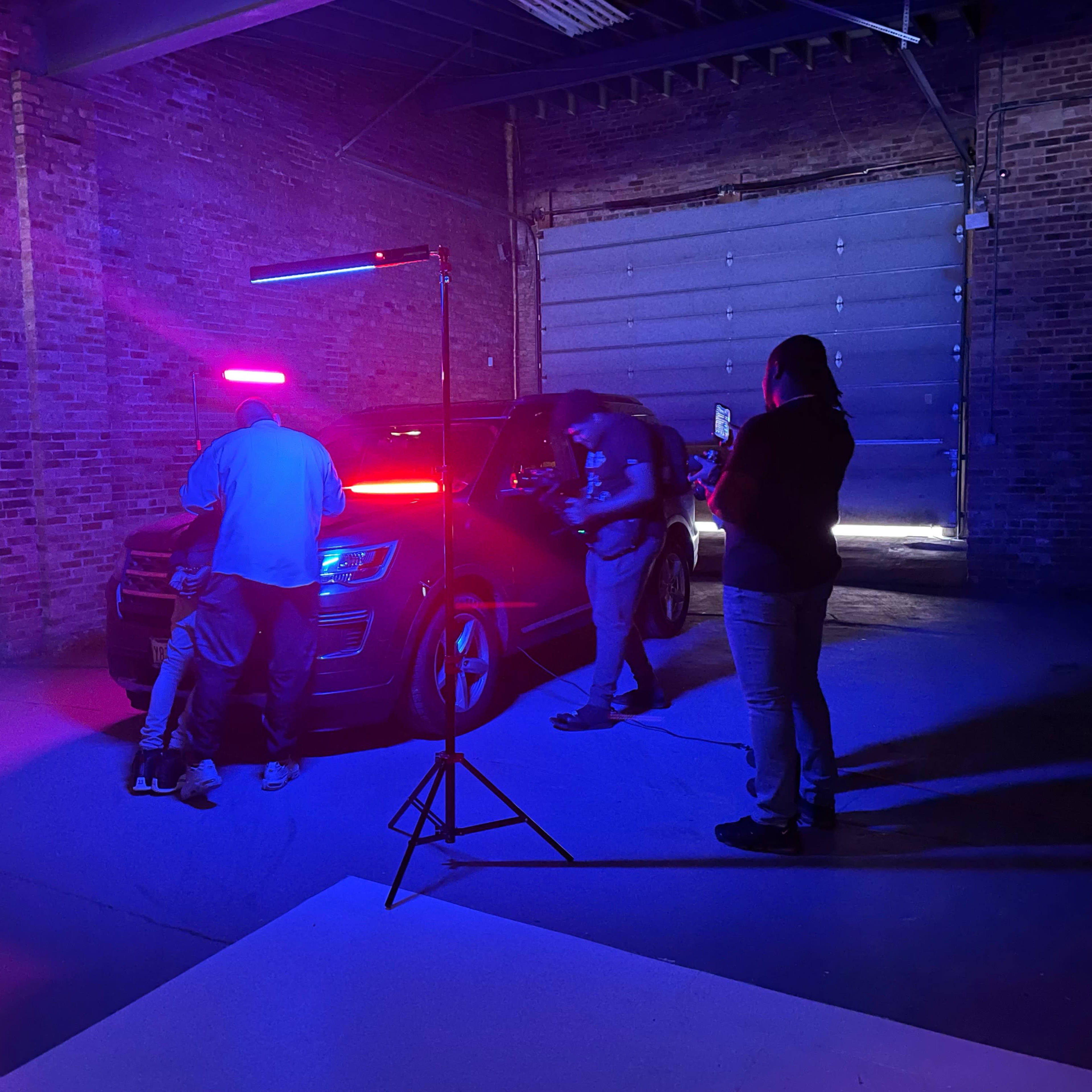 A group of people is setting up lighting equipment around a black vehicle in an industrial space with brick walls.