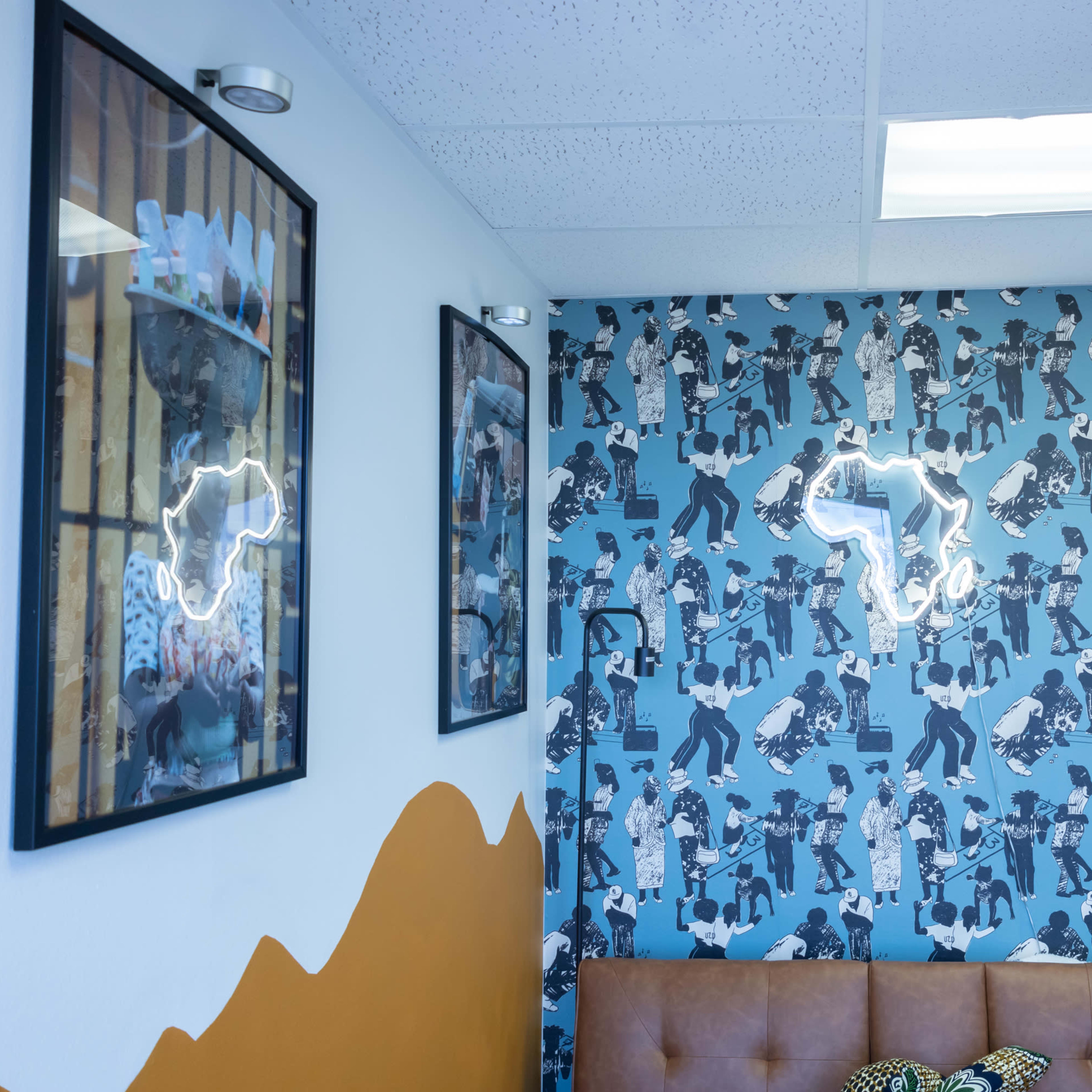 A corner of a room featuring patterned wallpaper with a light fixture in the shape of Africa, framed artwork on the walls, and a brown sofa.