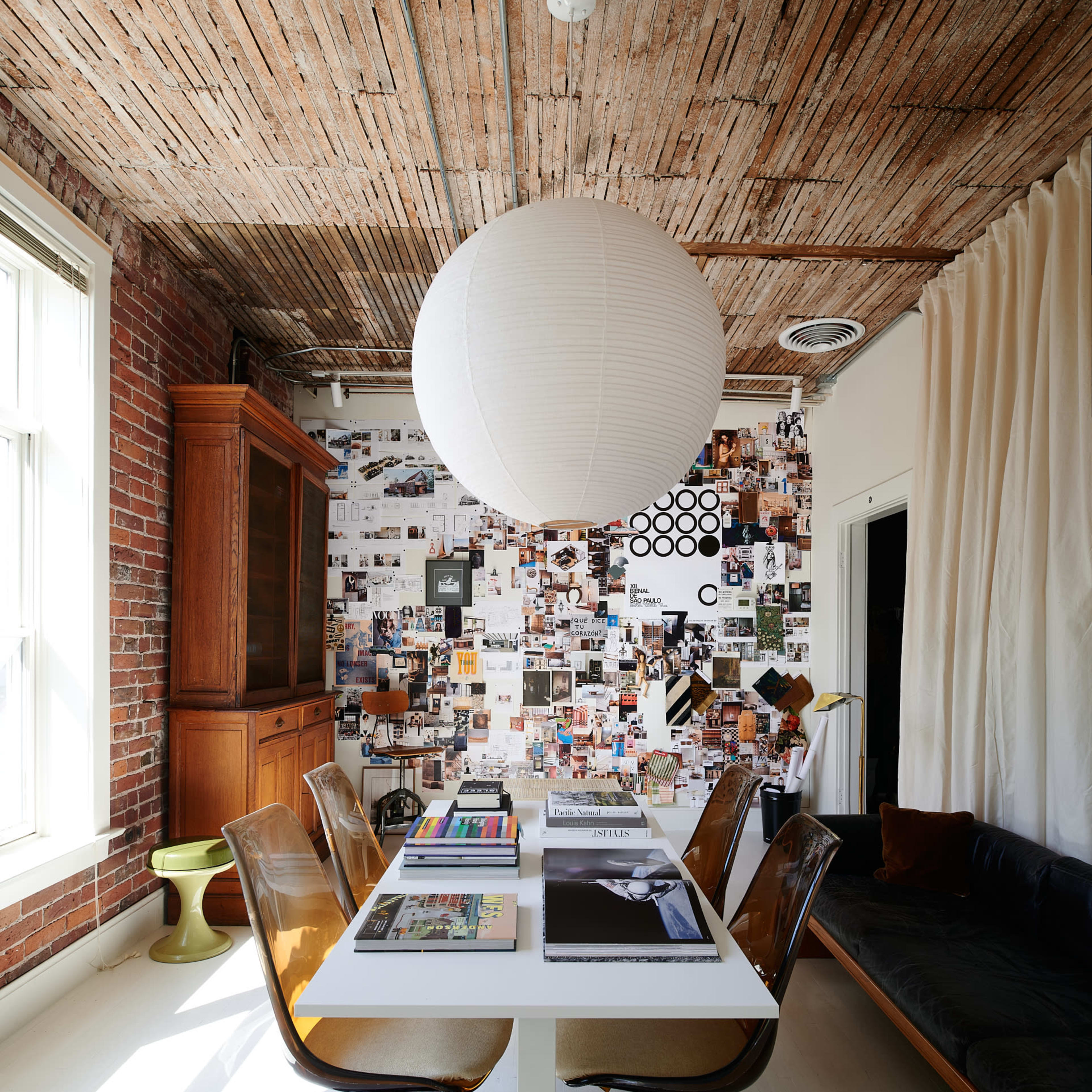 A dining area with a central table, surrounded by chairs, a wall covered in photographs and art, and a large paper lantern hanging overhead.
