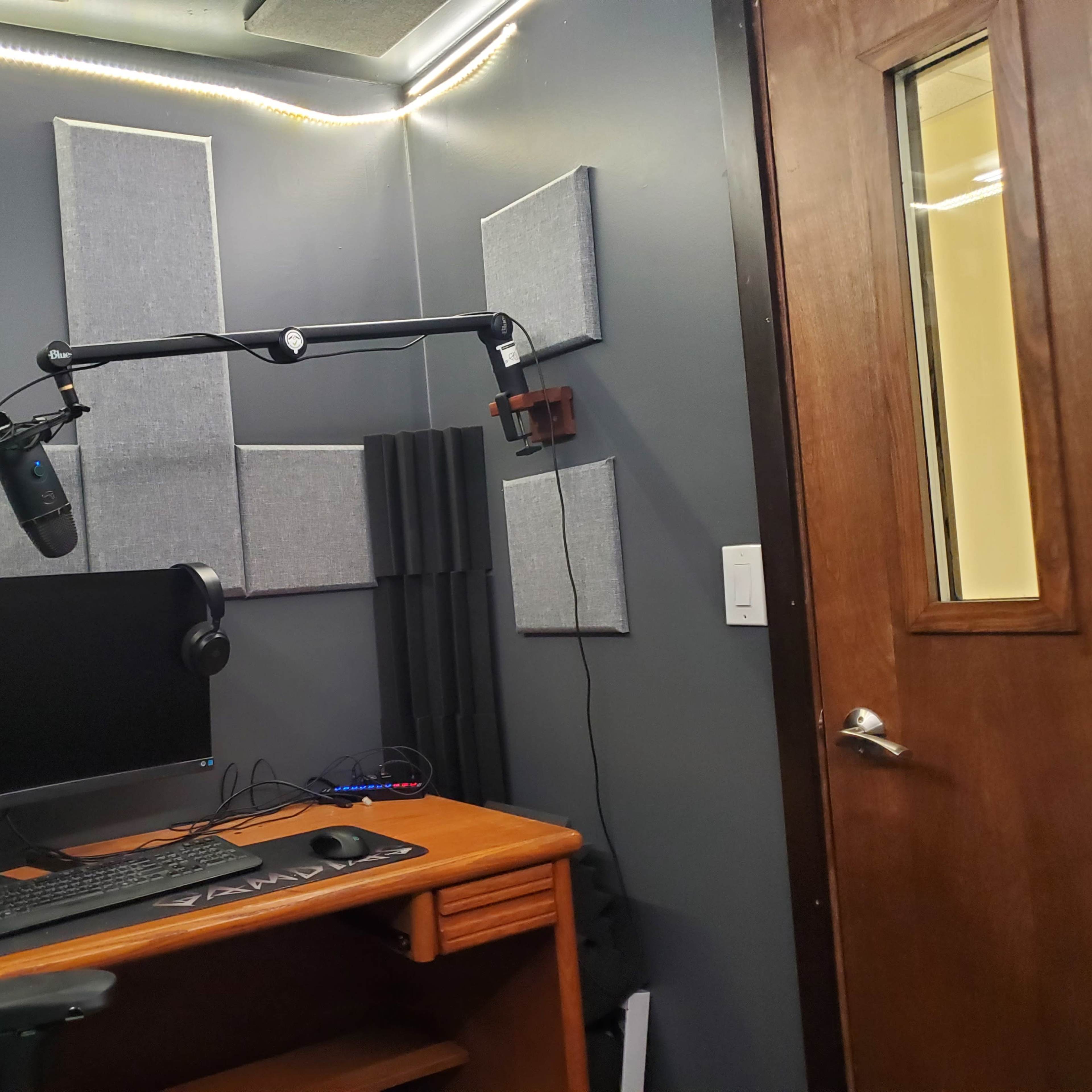 A recording studio setup with a computer monitor, microphone on an adjustable arm, headphones, and soundproofing panels on the walls.