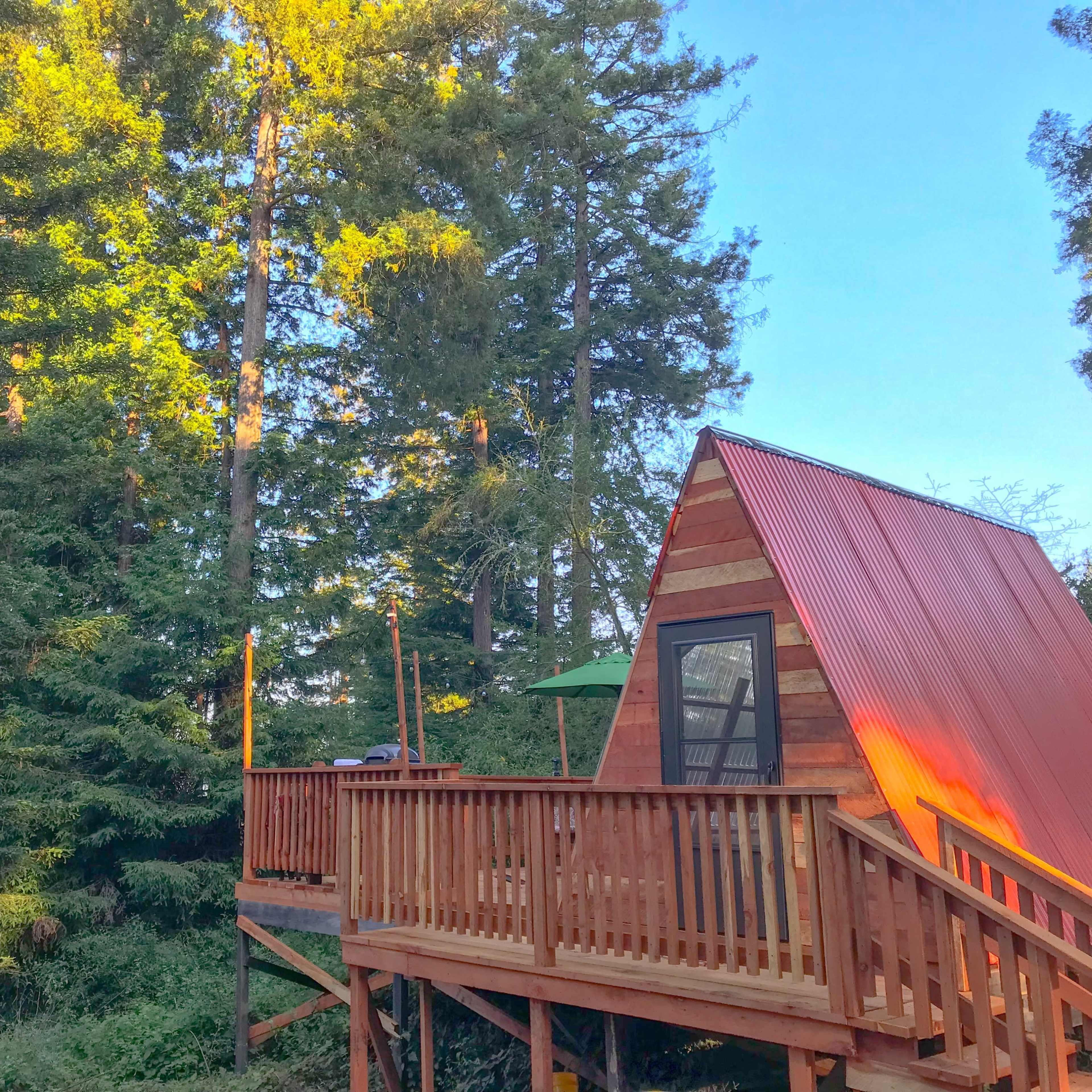 A wooden A-frame cabin with a red metal roof is nestled among tall green trees on a raised deck.