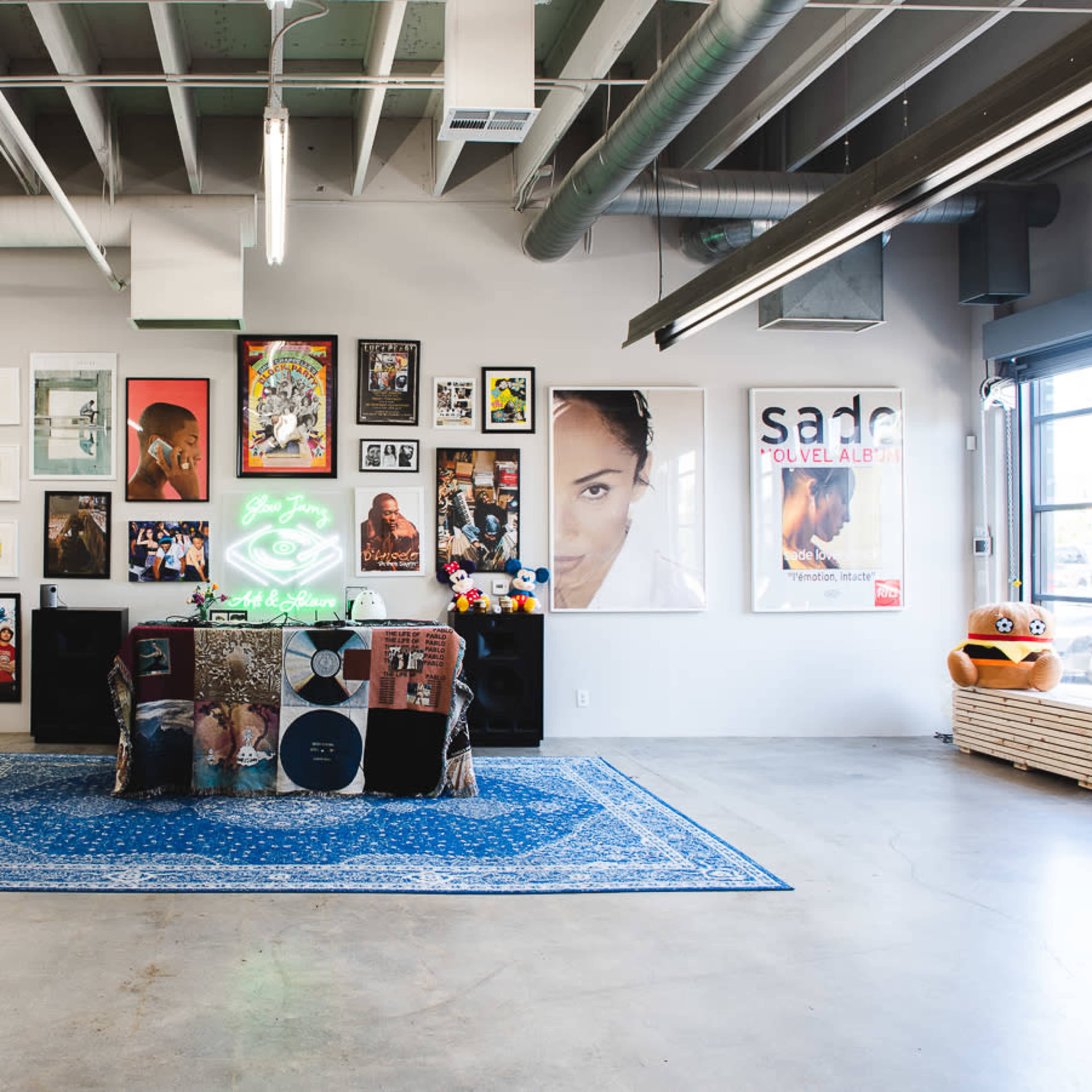 An art-filled gallery space with colorful artwork on the walls, a vintage rug on the concrete floor, and a neon sign in the center.