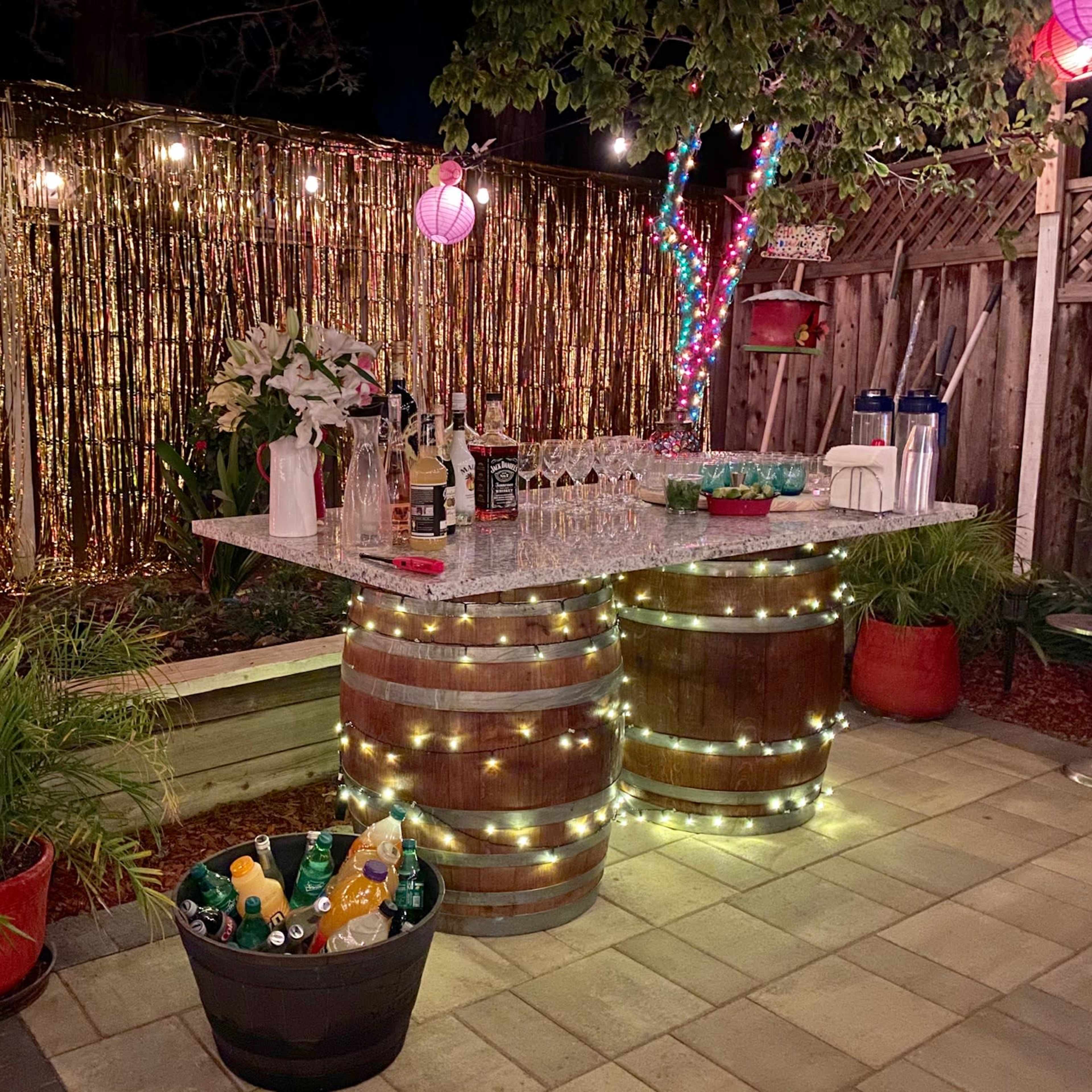 A backyard bar setup with a table made from barrels, decorated with lights and surrounded by tropical plants, under string lights and colorful lanterns.