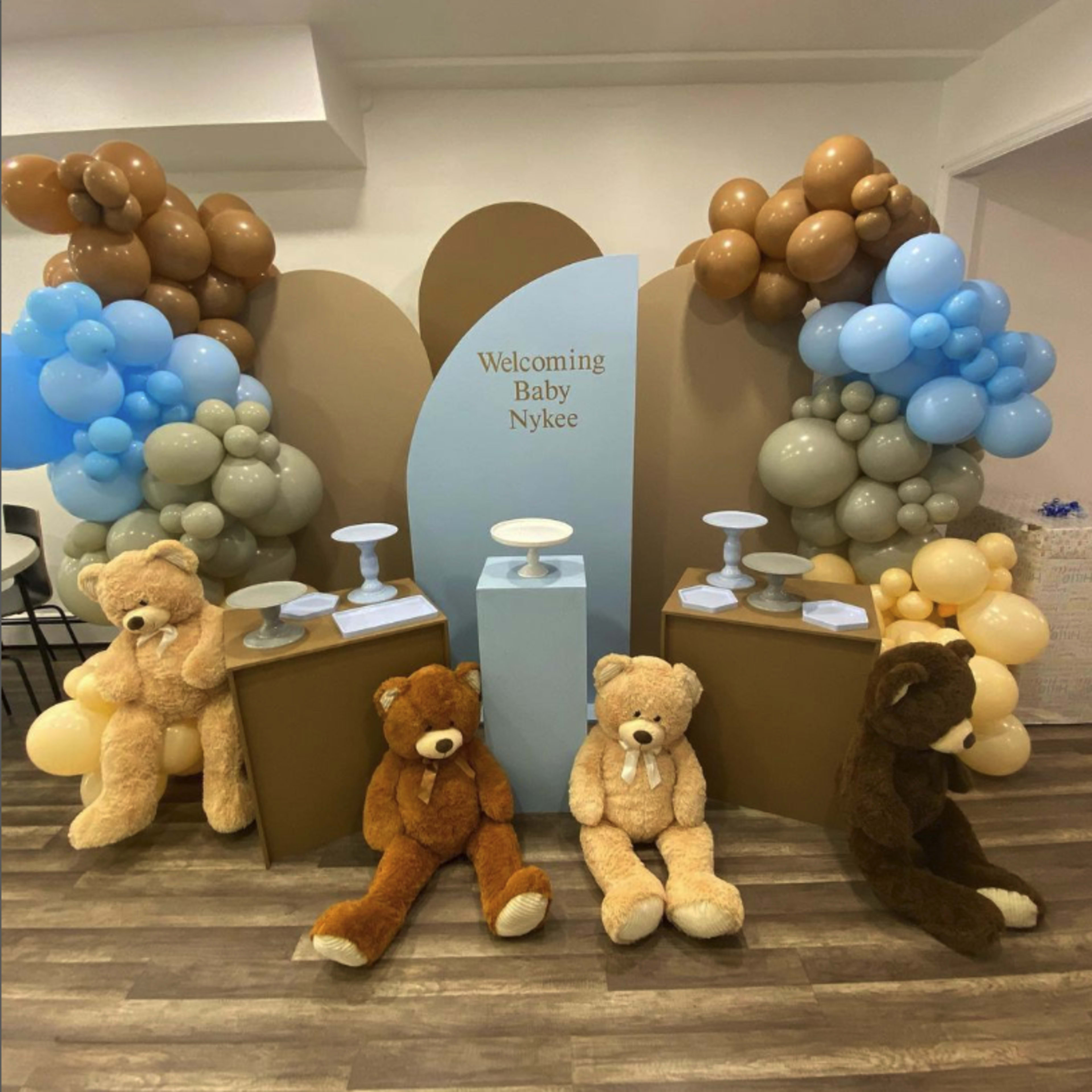 The scene features a decorated area for a baby welcoming event, with a backdrop of balloons in shades of blue, brown, and gray, flanked by large teddy bears sitting on the floor.
