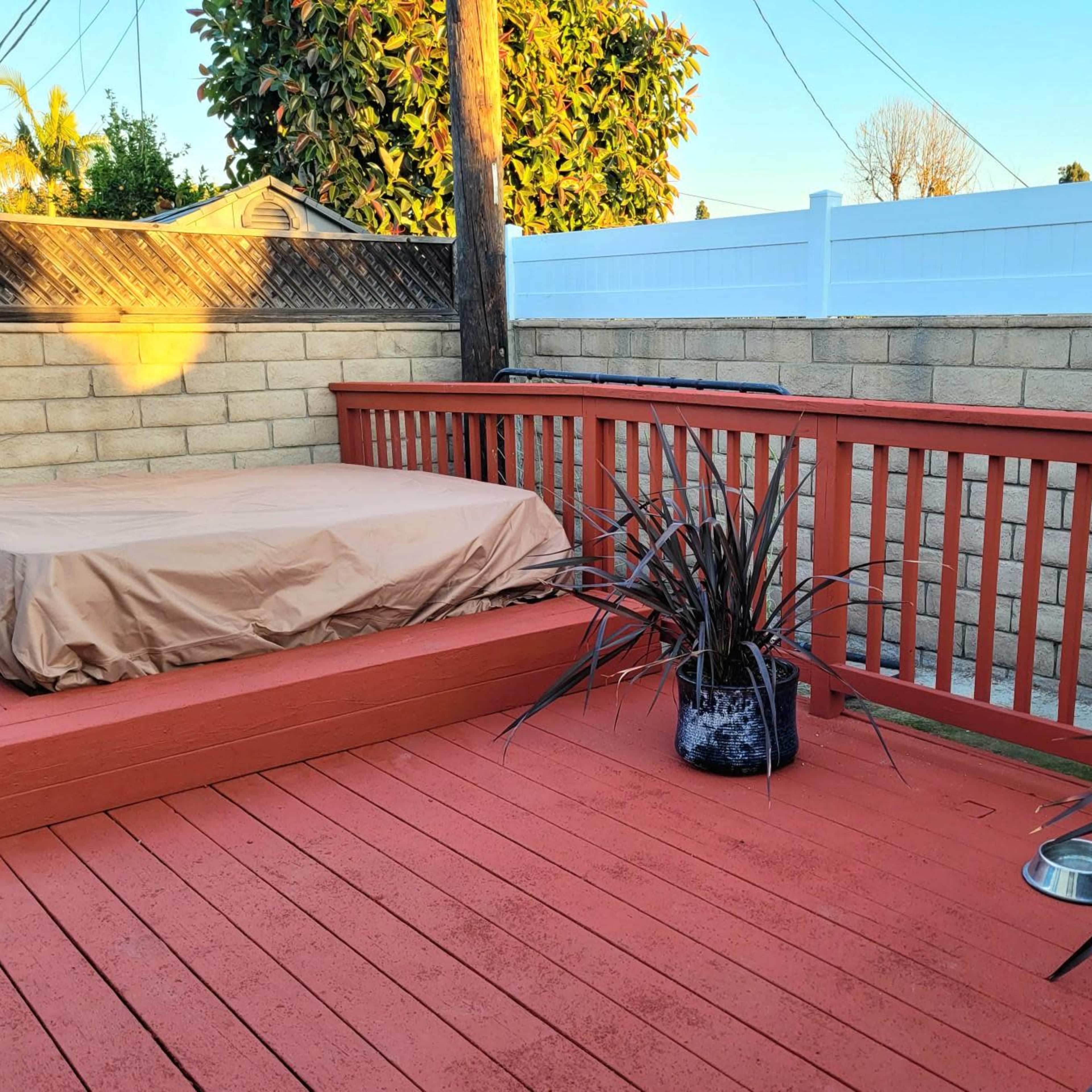 A wooden deck with a covered hot tub, potted plants, and a water bowl near a brick wall under a clear blue sky.