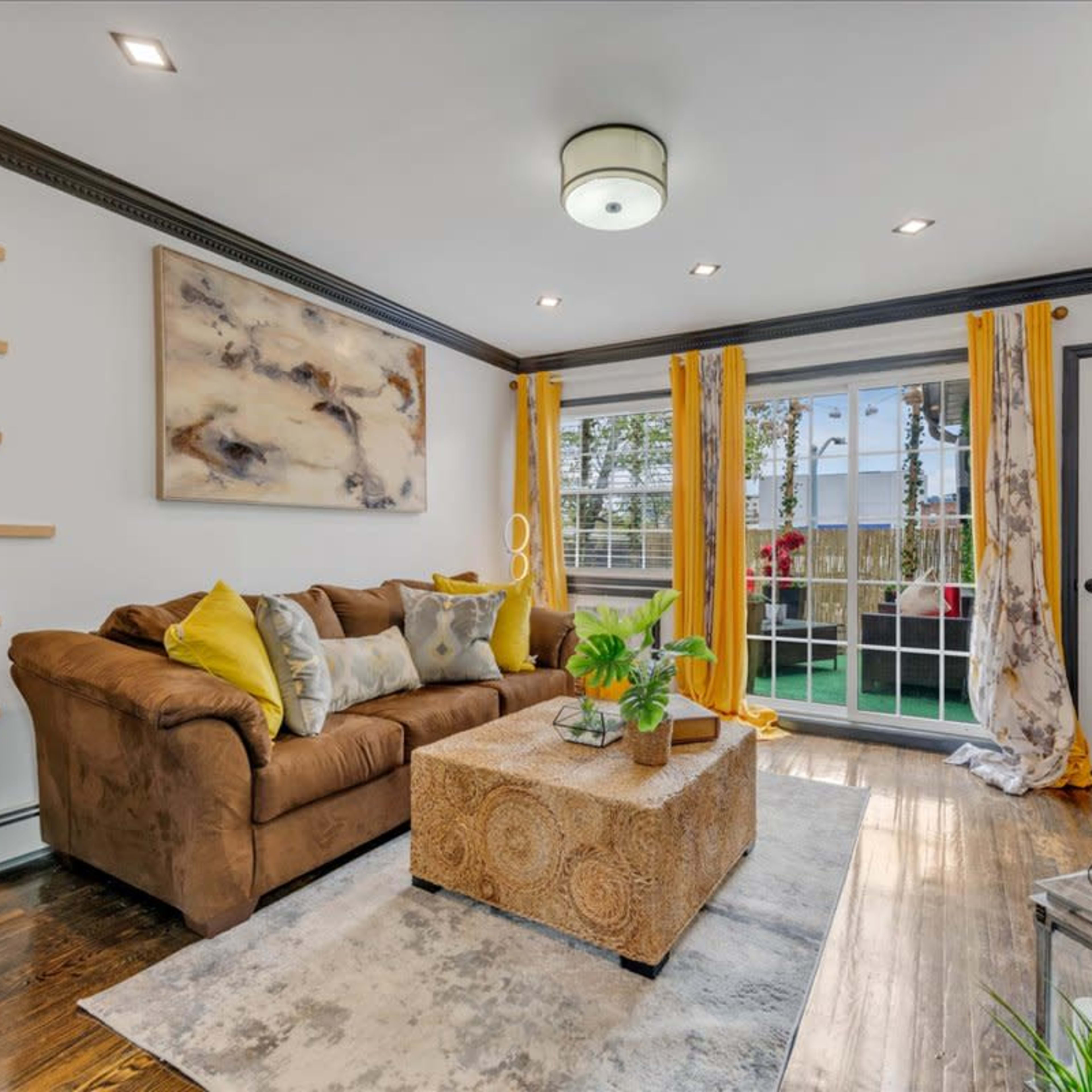 The living room features a brown couch with colorful pillows, a patterned coffee table, and large windows dressed with yellow curtains.