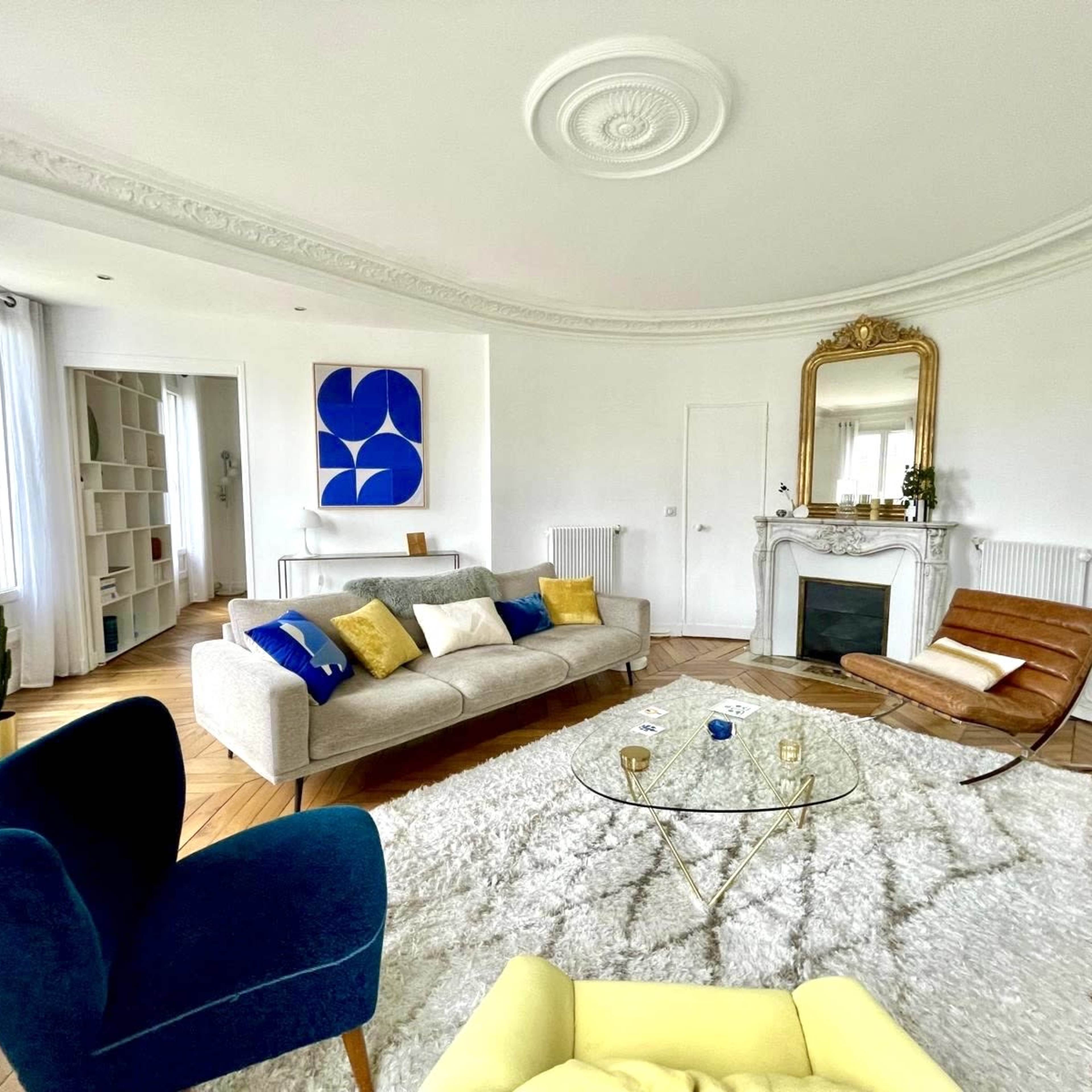 The living room features a stylish arrangement with a sofa, armchairs, a coffee table on a rug, and a decorative fireplace.