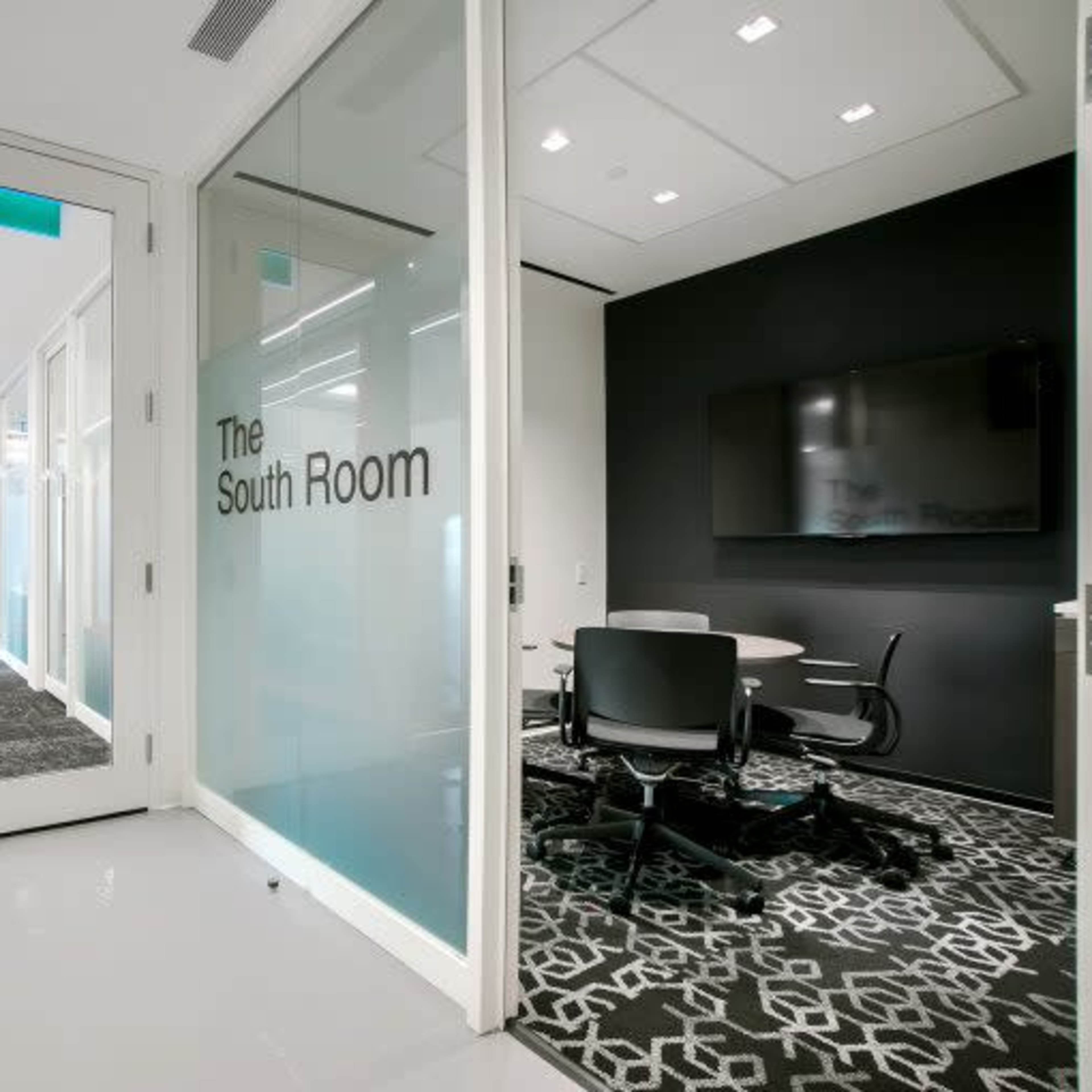 A modern office space with a glass-walled meeting room labeled "The South Room" and a dark wall featuring a wall-mounted screen.
