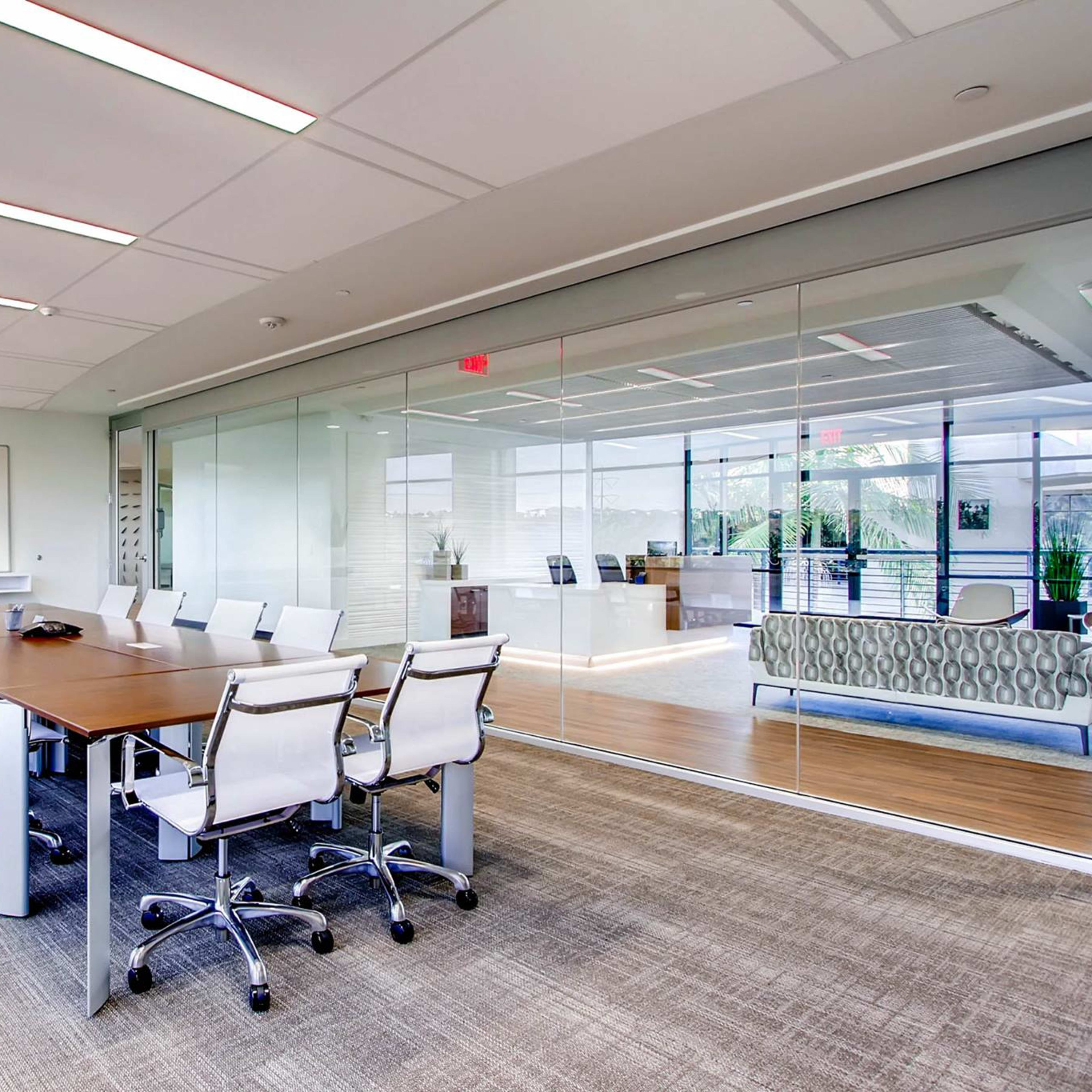 A large conference room features a long wooden table surrounded by white chairs, with glass walls and a view of an office space.