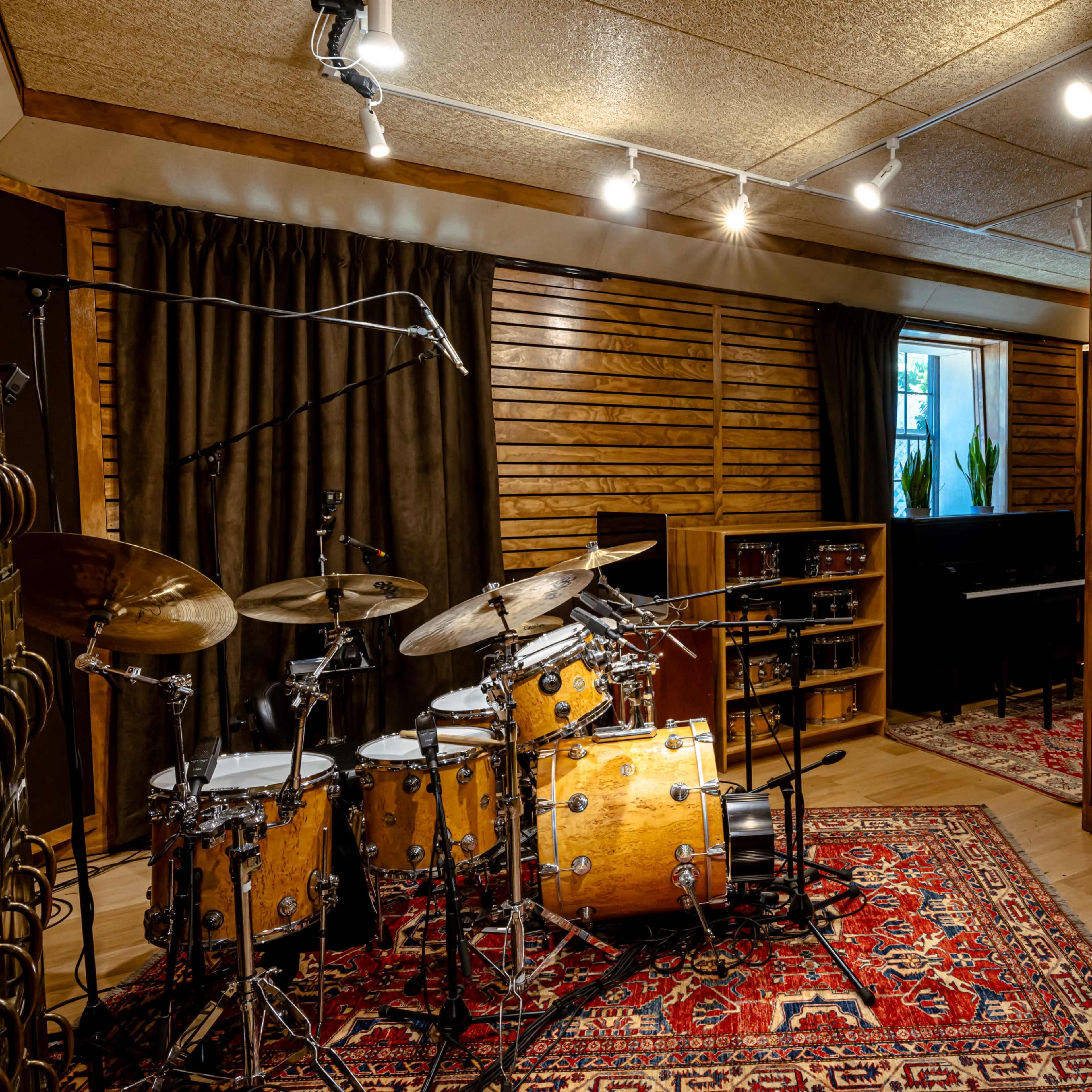 A professional music studio featuring a drum set, a piano, and soundproofed walls with warm wooden accents.