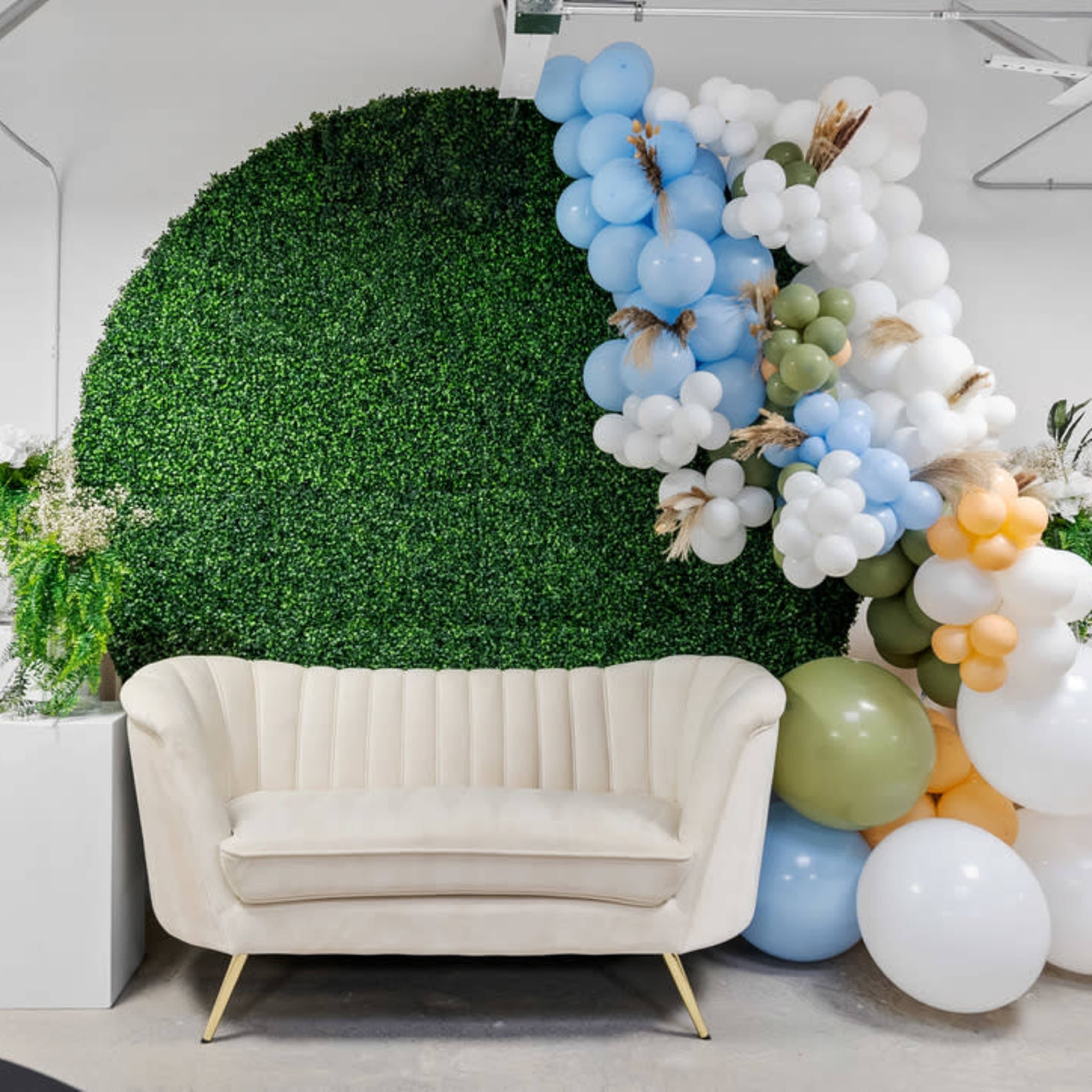 A beige sofa is positioned in front of a green wall of artificial foliage, accented by a cluster of balloons in various colors.