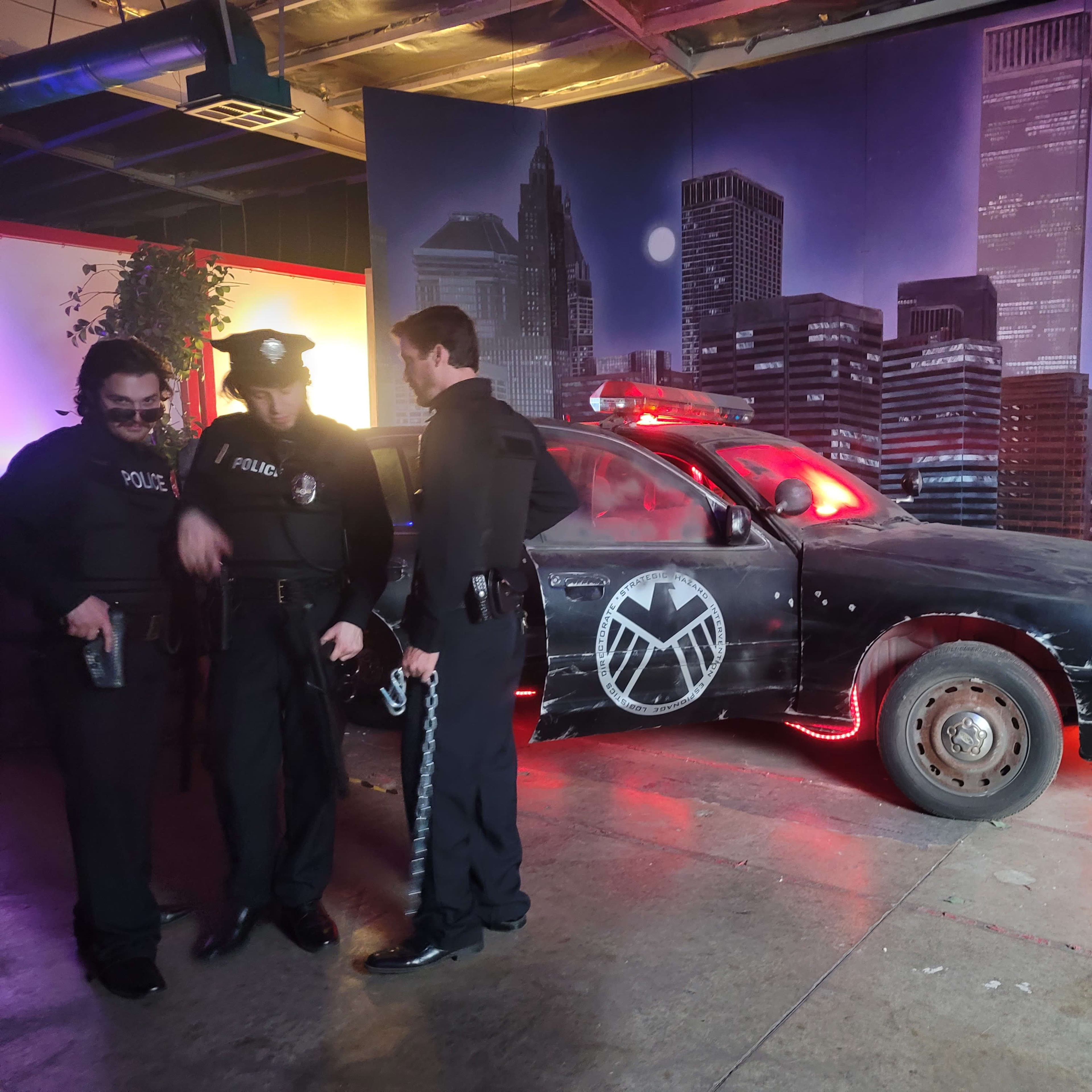 Two police officers stand beside a police car featuring a large emblem, set against a backdrop of a city skyline.