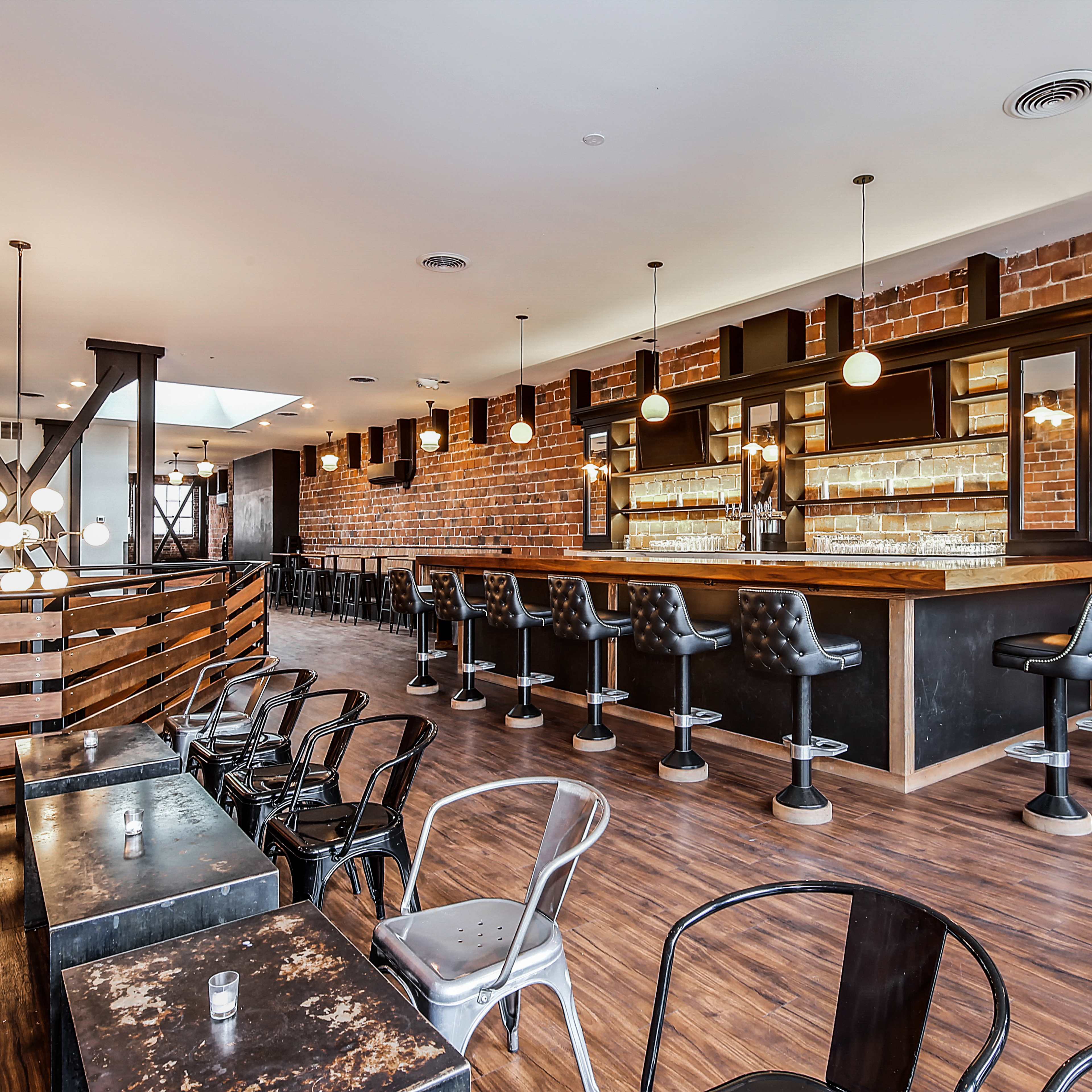 A modern bar with a brick wall, featuring black stools, a countertop, and tables in a spacious interior.