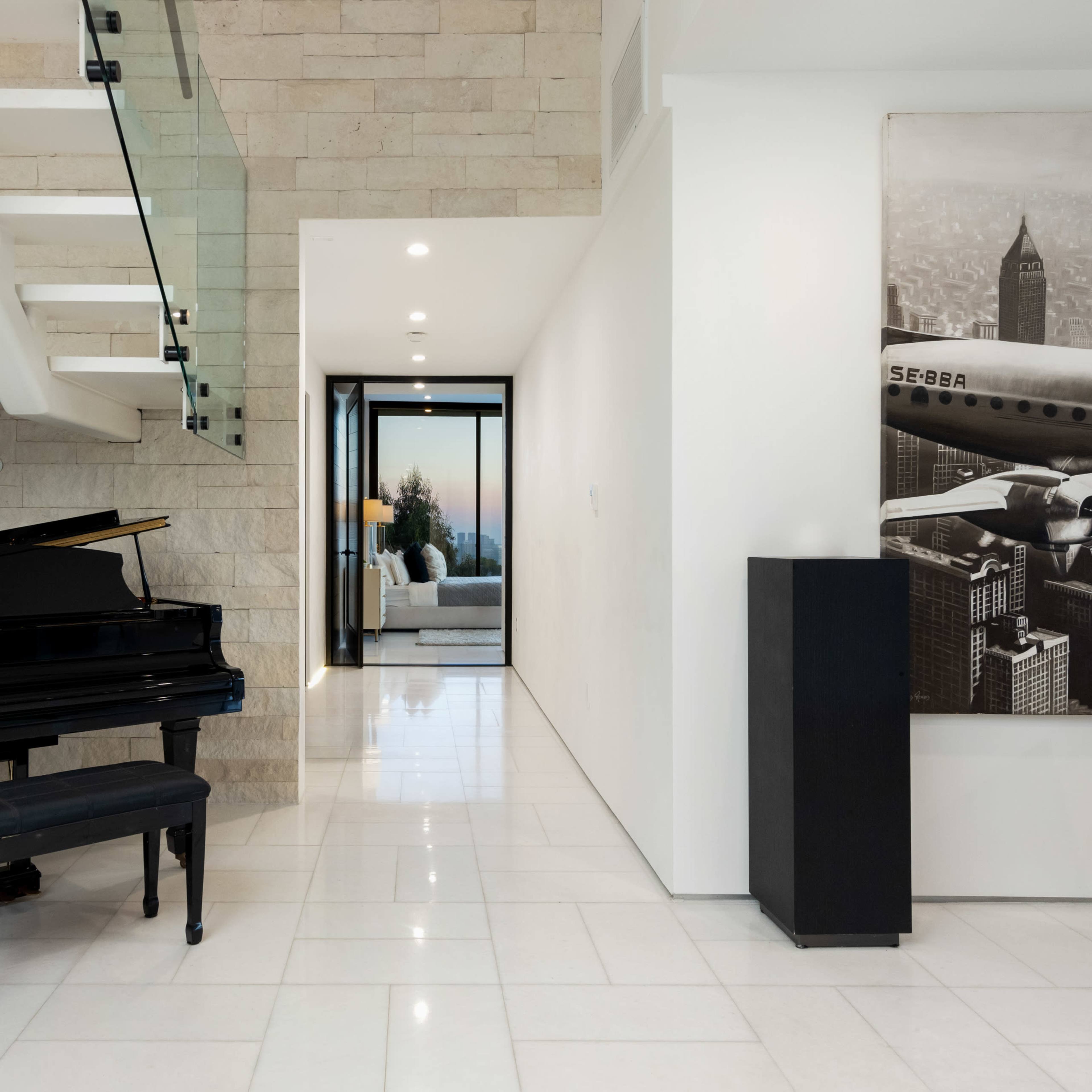 A black grand piano is positioned in a modern foyer with stone walls and a staircase leading to an upper level.