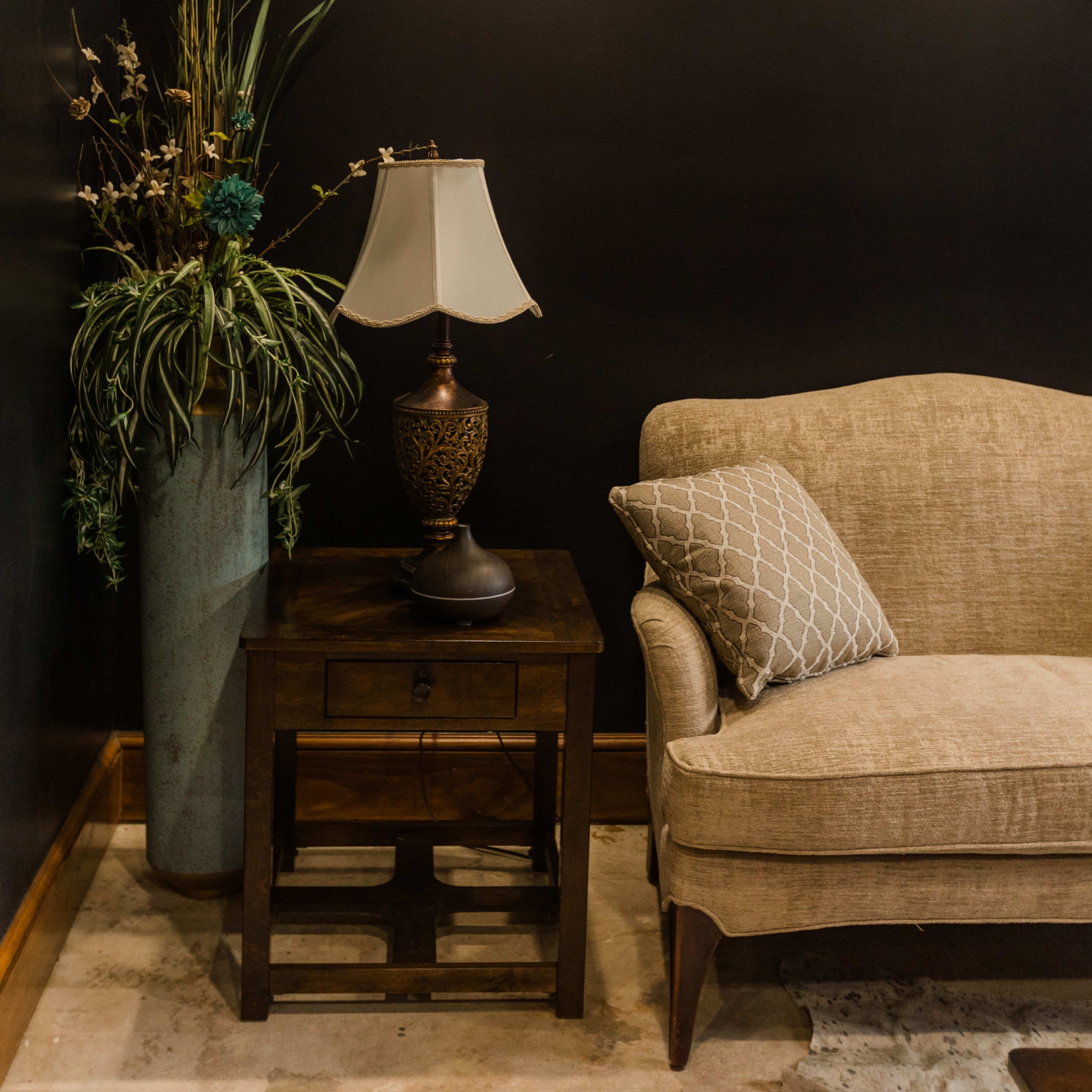 The scene features a brown sofa next to a wooden side table with a lamp, accompanied by a tall planter with decorative plants against a black wall.