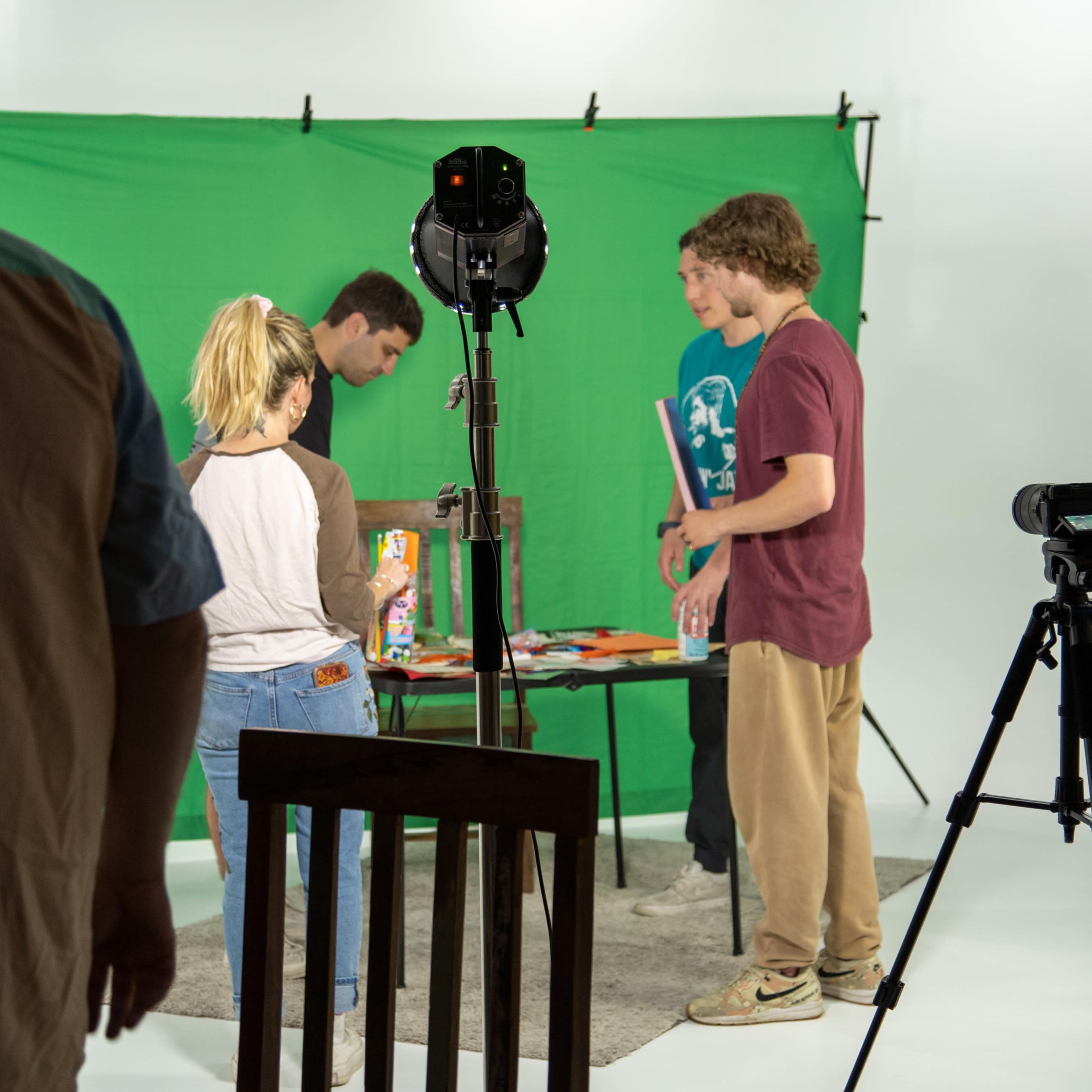 Five individuals are gathered around a table with various items in front of a green screen backdrop, while a camera and lighting equipment are set up nearby.