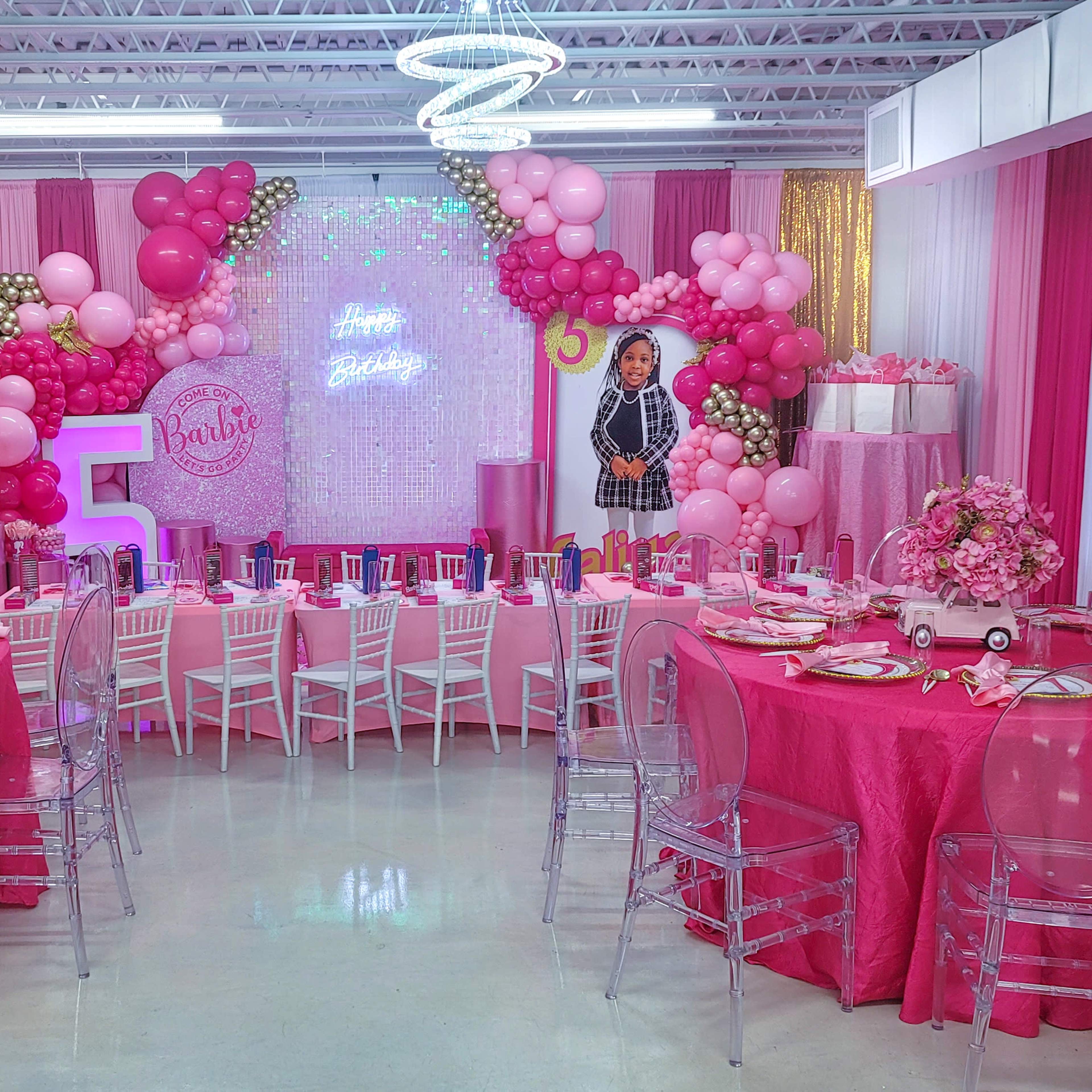 A brightly decorated birthday party scene with pink and white balloons, tables covered in pink tablecloths, and a large "5" centerpiece surrounded by themed decorations.