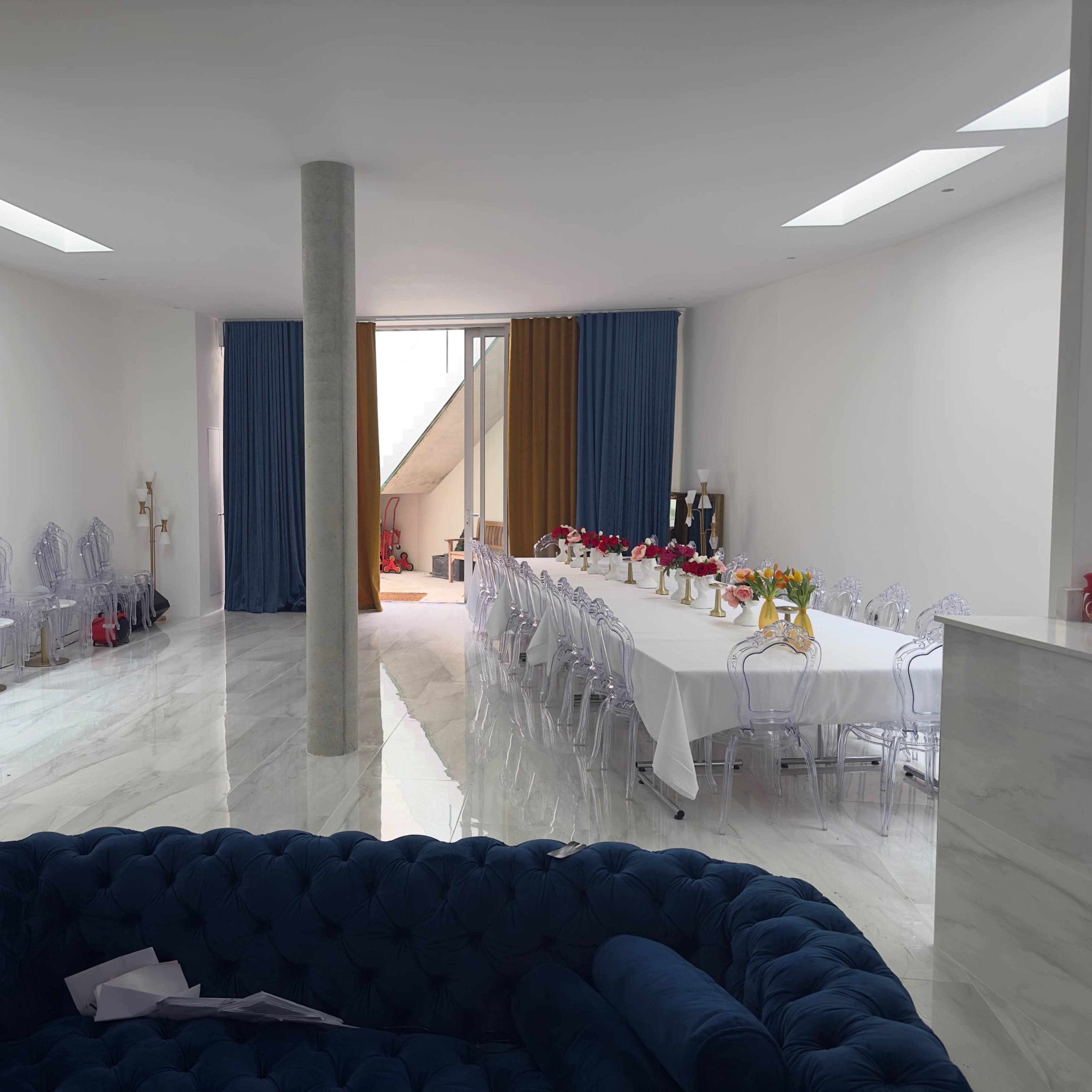 A modern dining area features a long white table adorned with flowers, surrounded by transparent chairs, and is illuminated by natural light from large windows.