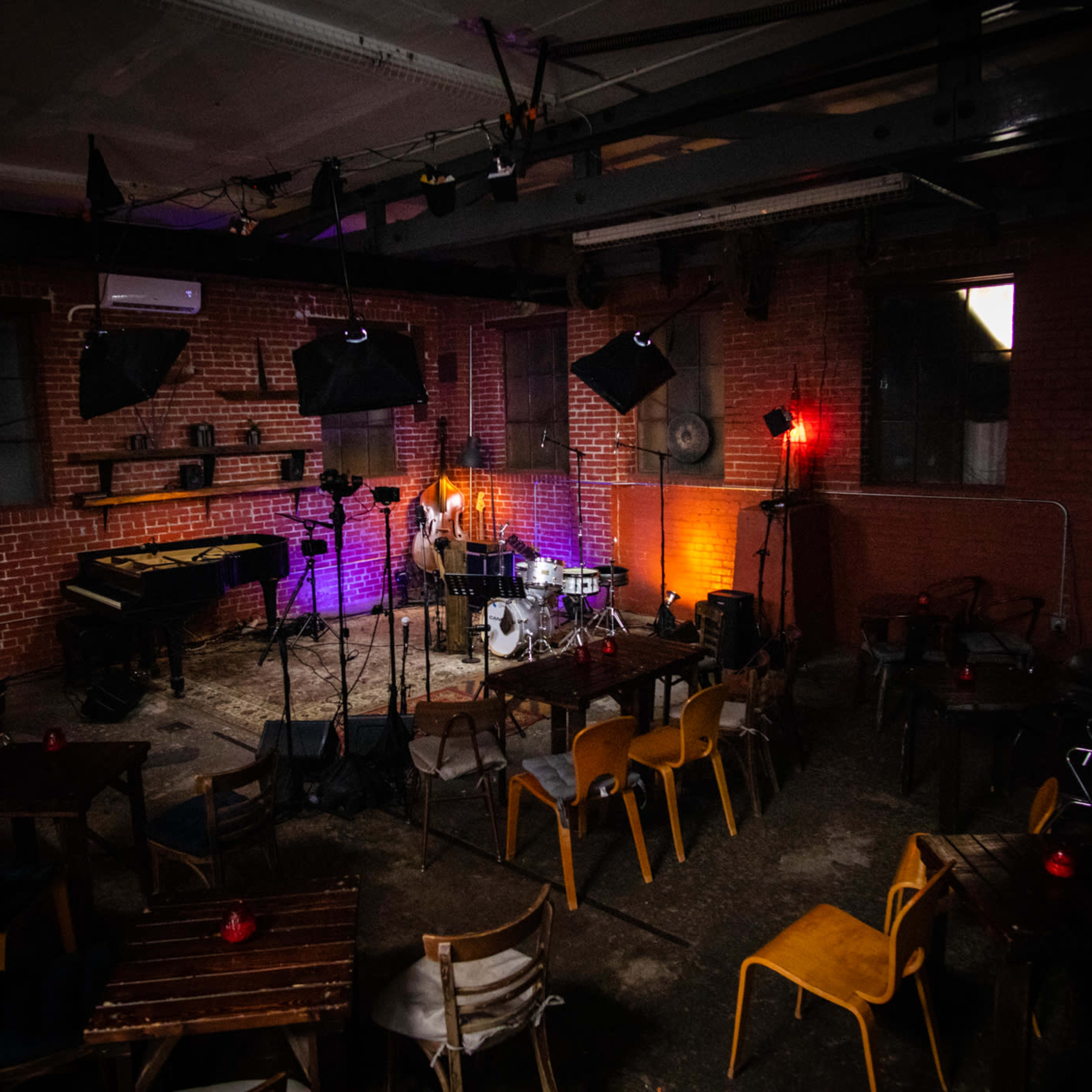 A dimly lit music venue with a stage set up for a performance, featuring a drum kit, microphone, and a piano, surrounded by empty chairs and tables.