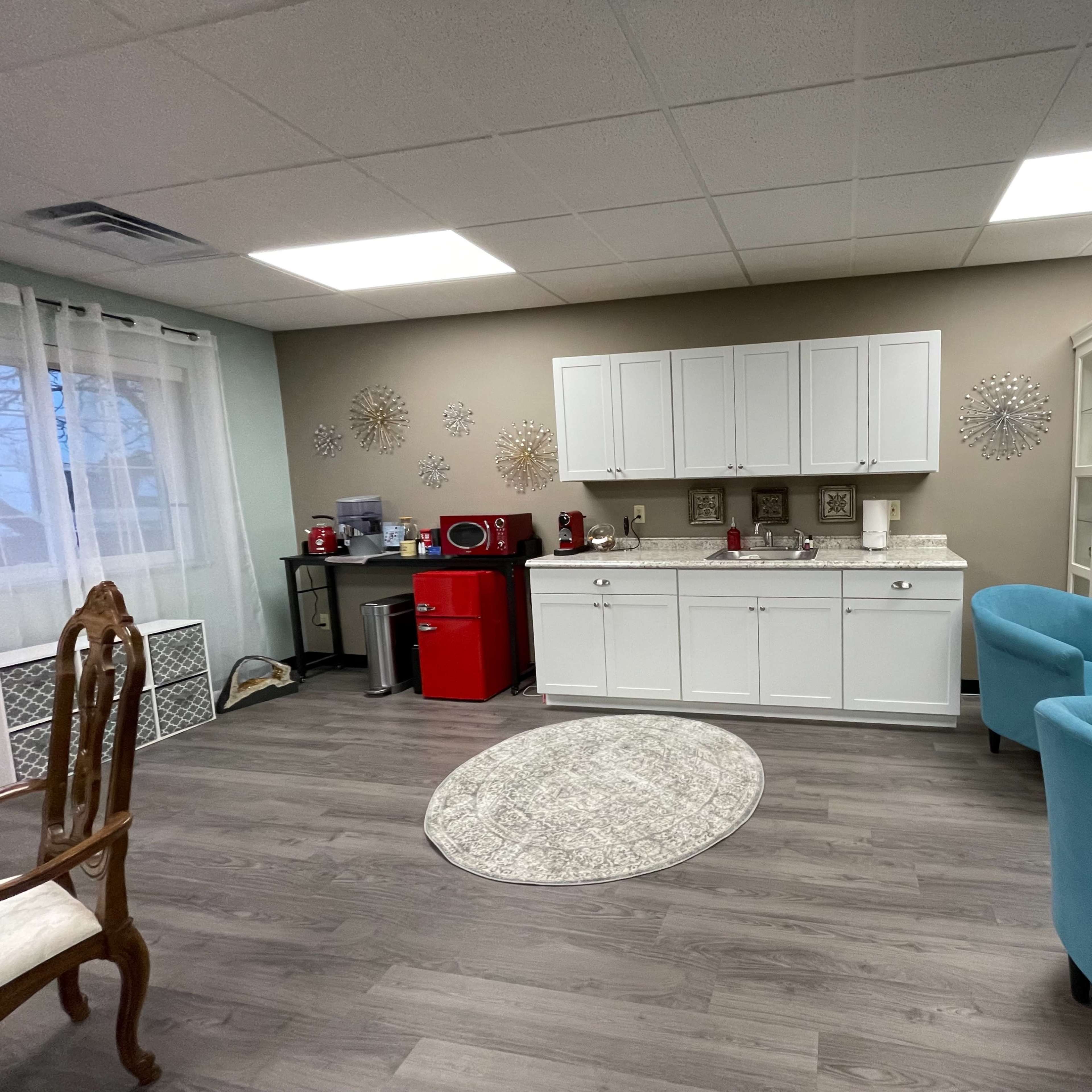 A well-lit room featuring a kitchenette with white cabinets, a red mini refrigerator, and a display cabinet, along with light blue chairs and a round area rug.